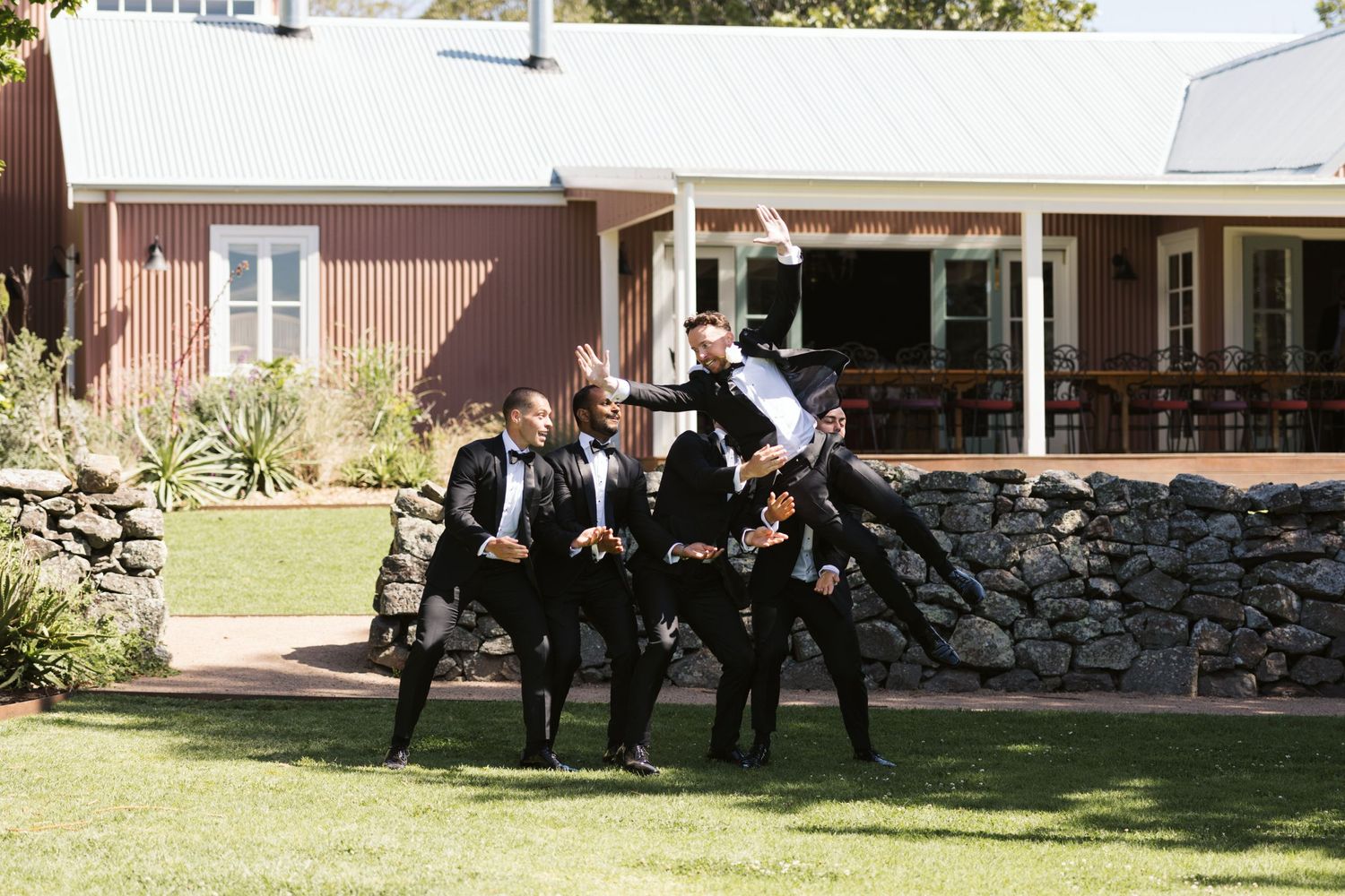 Wedding party members gather in front of a brown house with stone wall and white trim.