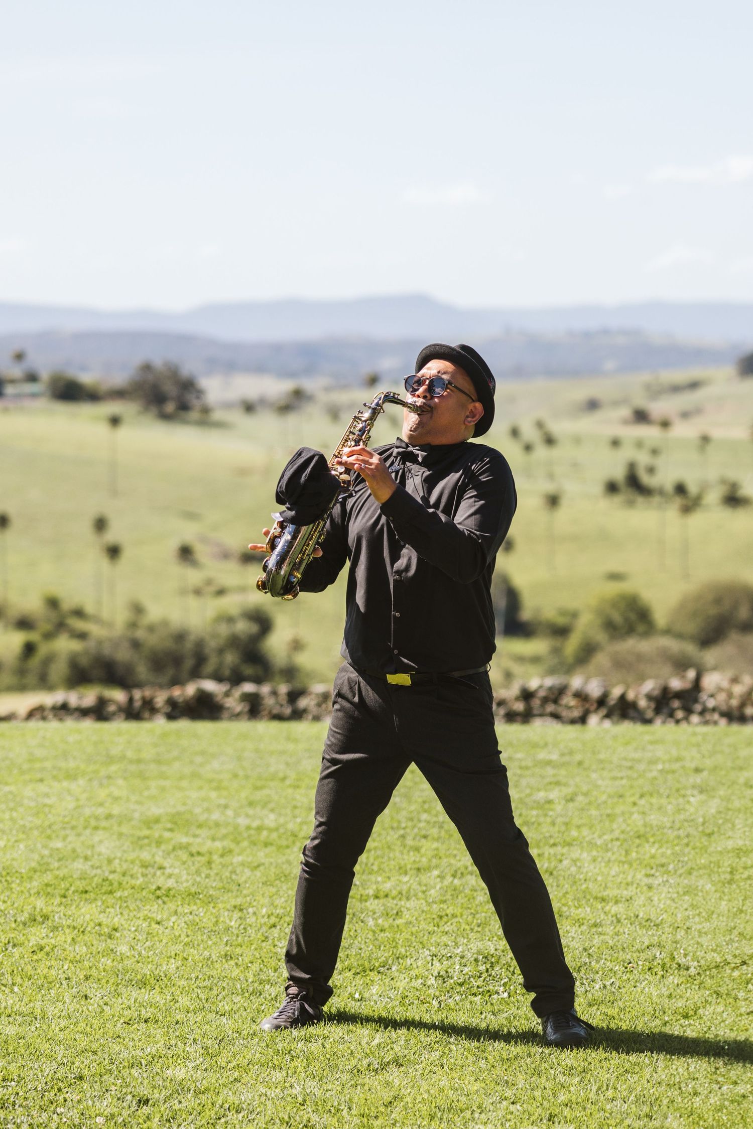 Musician in dark attire playing saxophone on a grassy hilltop overlooking a scenic mountain landscape.
