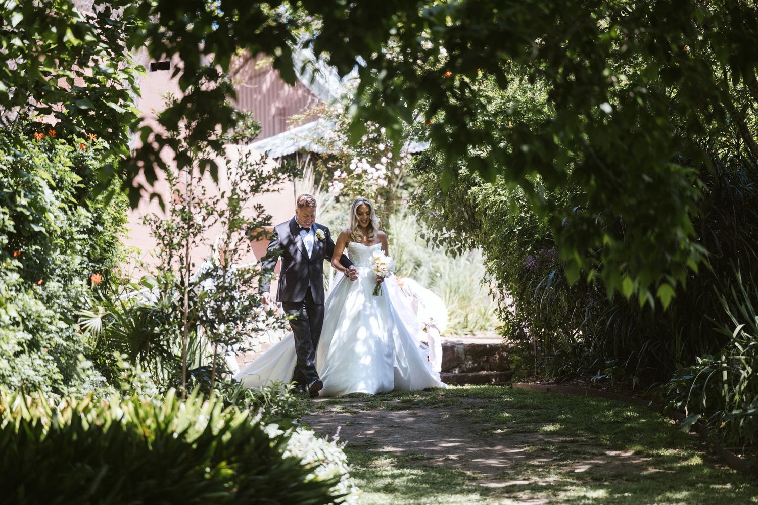 Newlywed couple walks along garden path under tree canopy.
