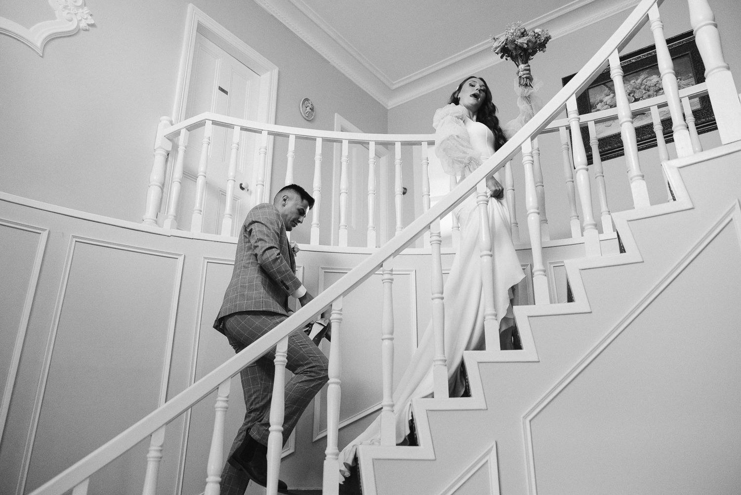 Black and white wedding photo of bride and groom on elegant staircase, bride holding bouquet aloft.