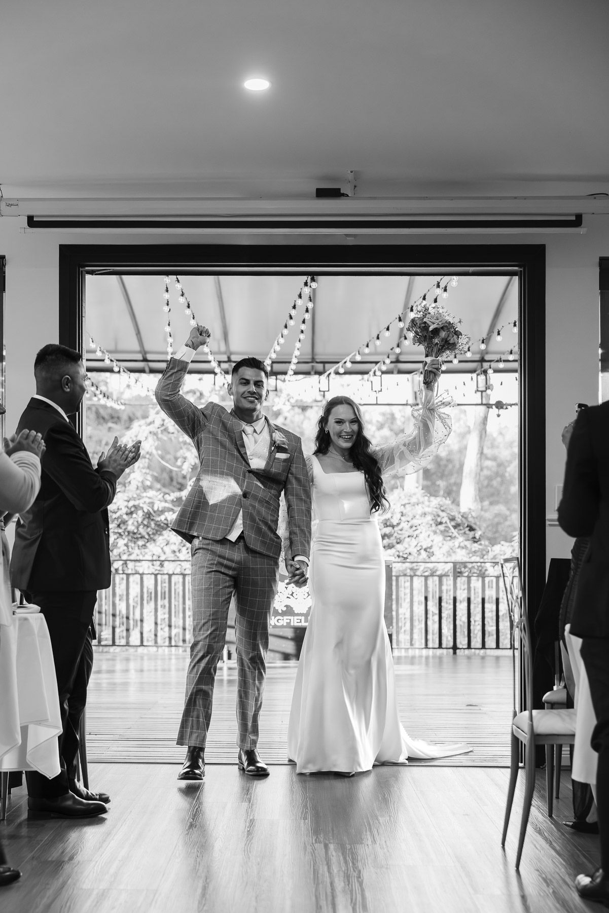 Black and white photo of a joyful bride and groom raising their arms as they enter their wedding reception.