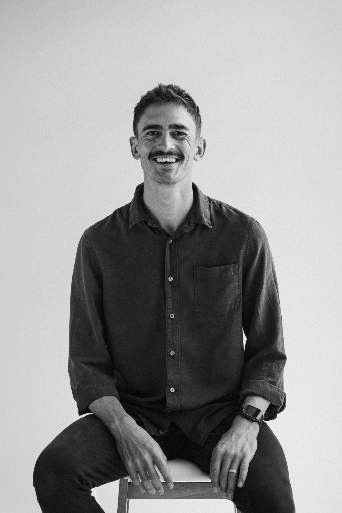 Black and white portrait of a smiling young man with mustache sitting on a stool wearing a button-up shirt.