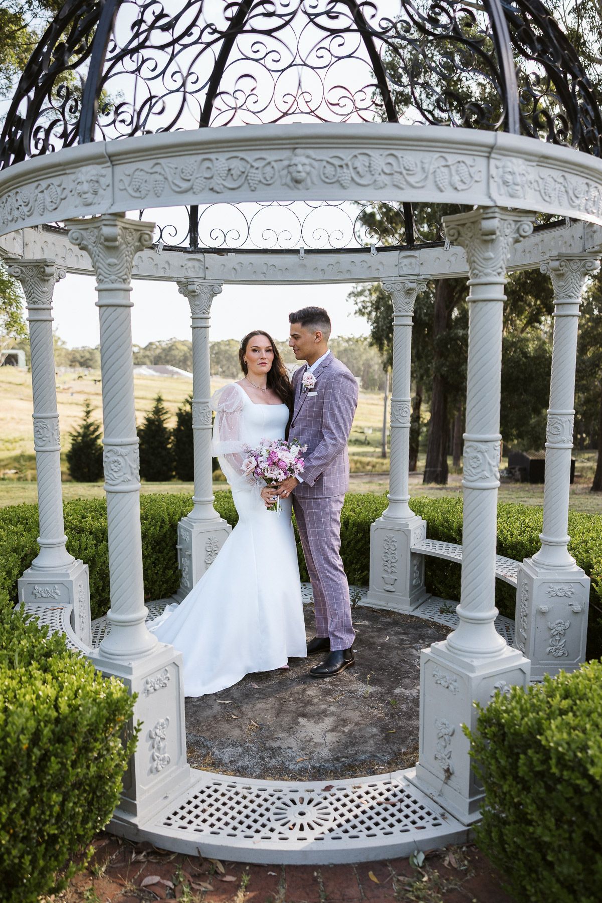 Bride and groom pose romantically under an elegant white gazebo surrounded by lush greenery on their wedding day.