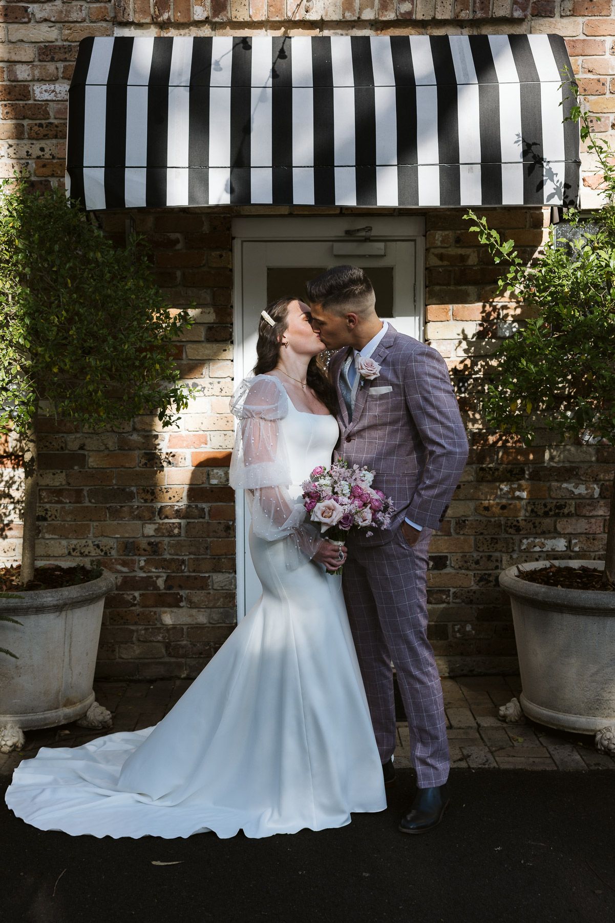 A bride and groom share a kiss outside a brick building with a striped awning, bride holding a pink bouquet.