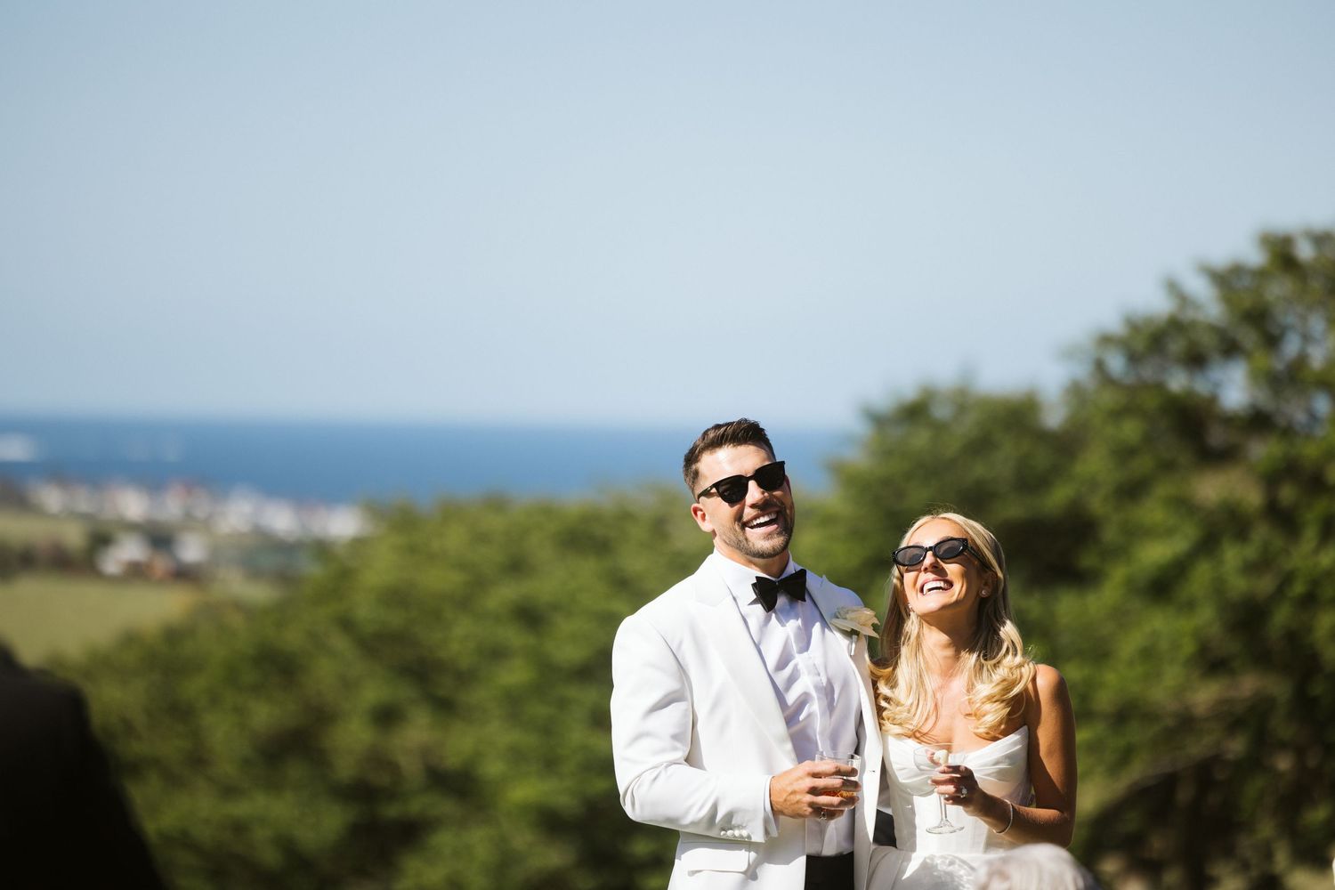Joyful wedding couple sharing laughs on a hilltop with ocean views in the background.