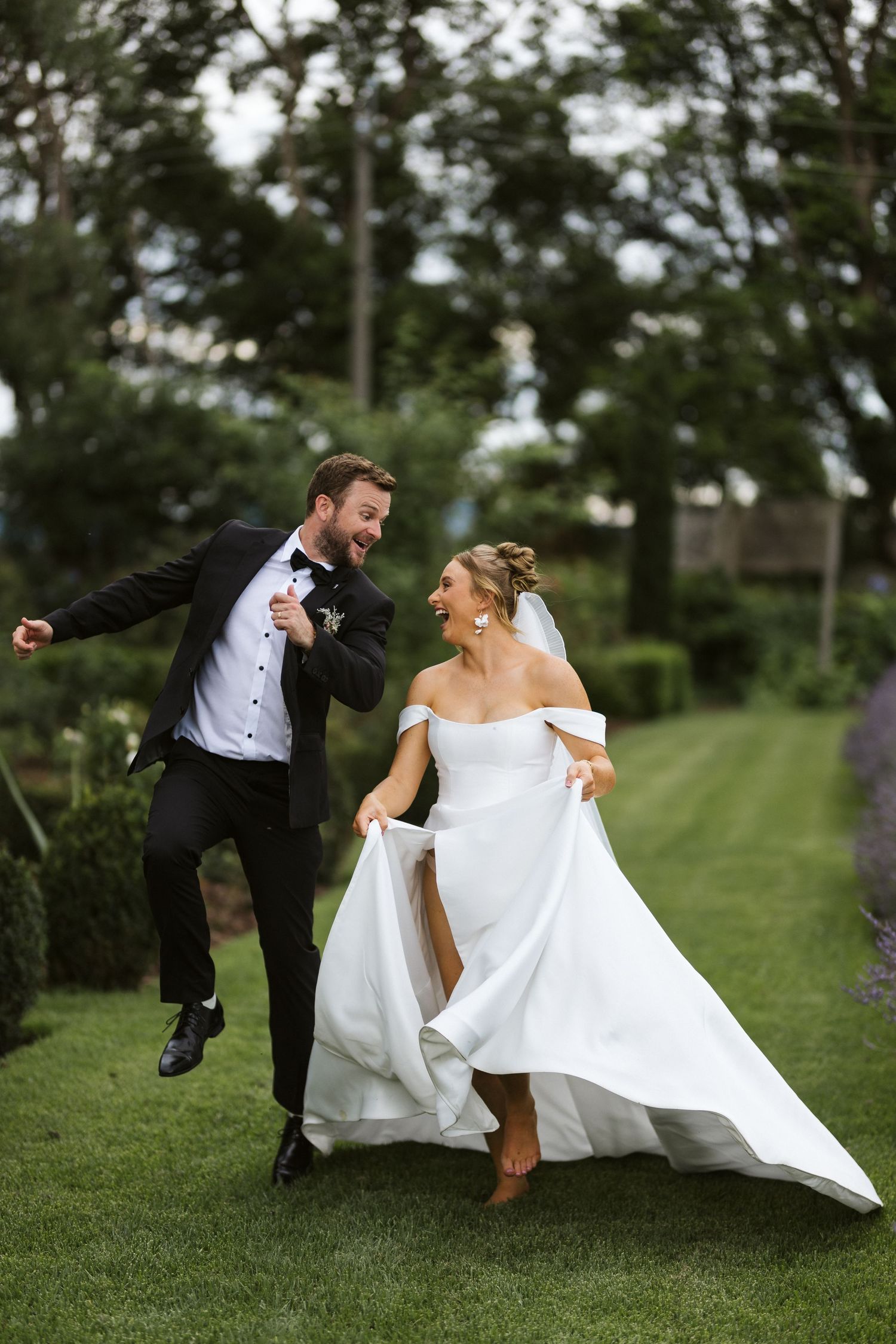 Energetic wedding couple dances and jumps playfully on garden lawn in formal attire.