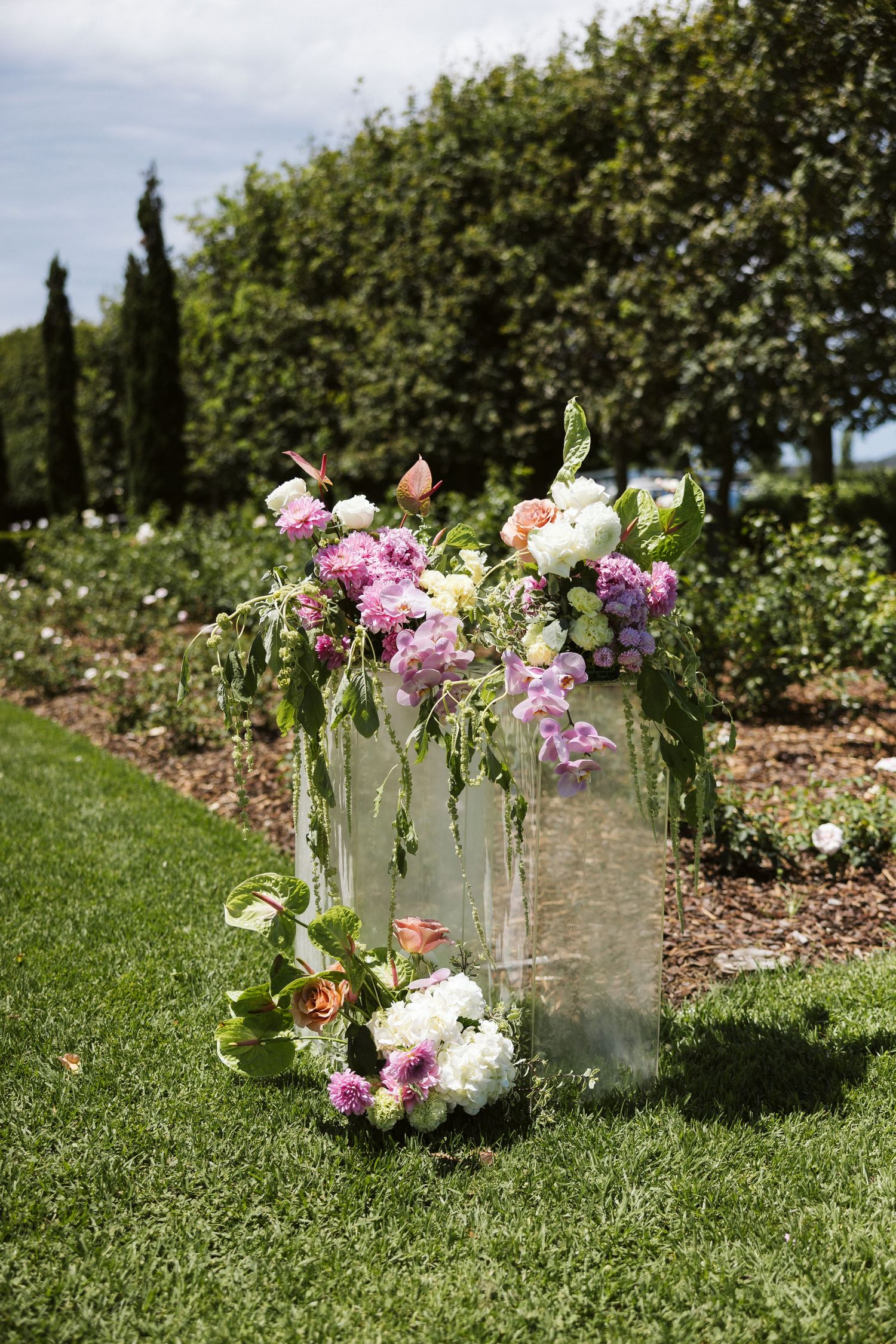 Elegant floral arrangement in glass vases with pink and purple flowers against a garden backdrop with cypress trees.