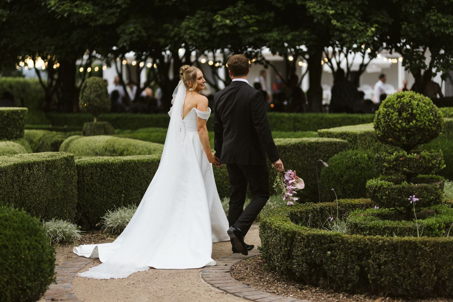 Newlyweds walking through manicured garden hedge maze on wedding day.