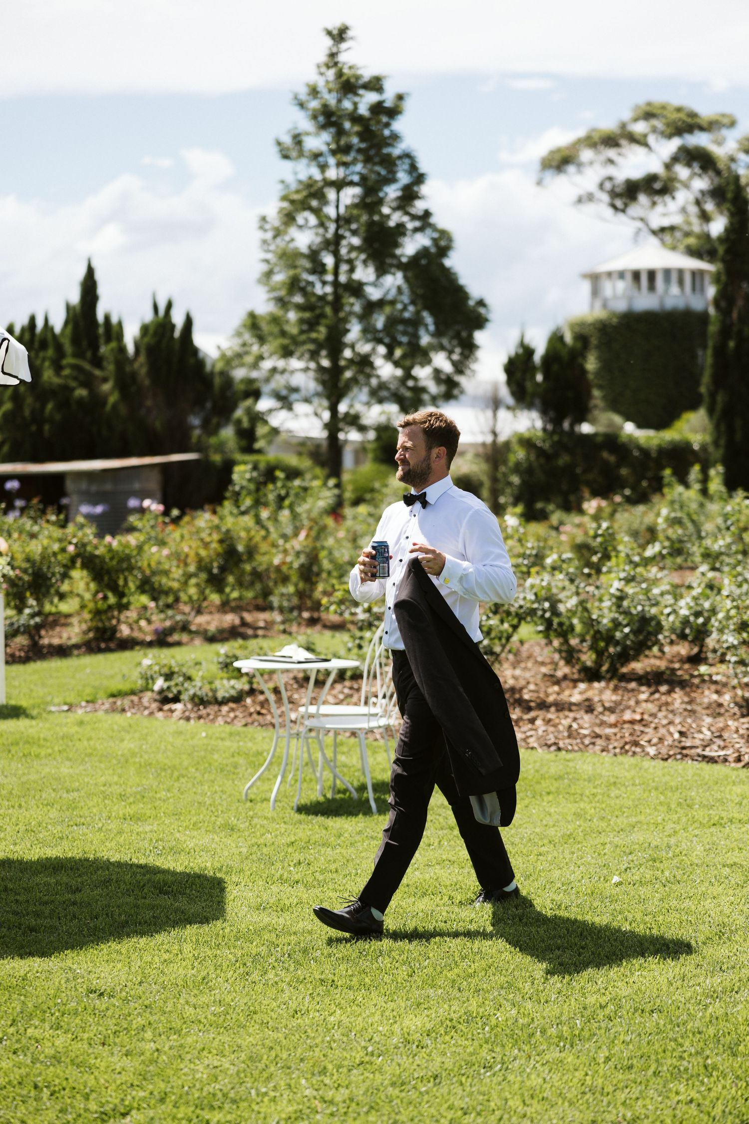 Formal-dressed attendant carrying jacket while walking across garden lawn.