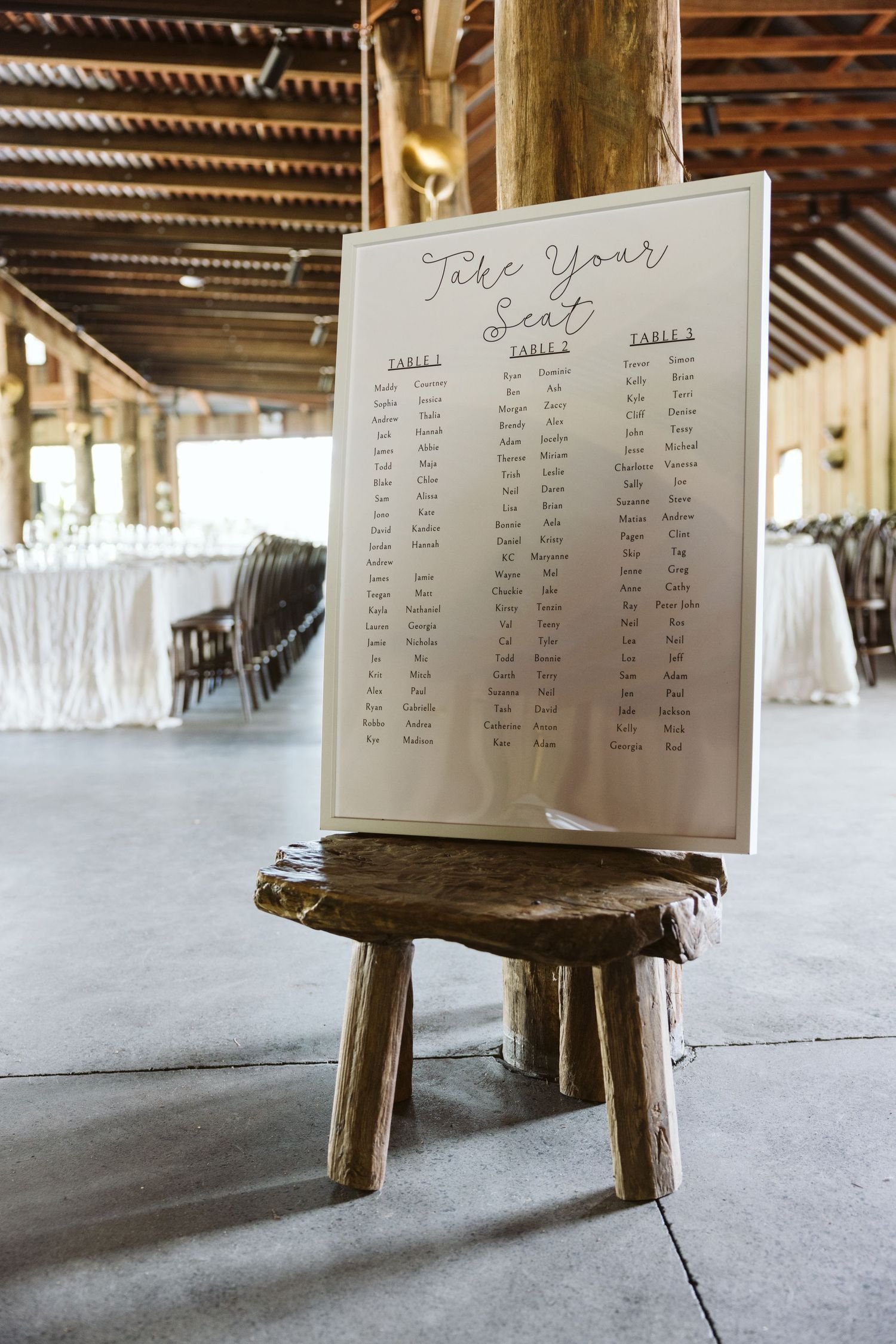 Wedding seating chart displayed on rustic wooden stand in reception venue with string lights.