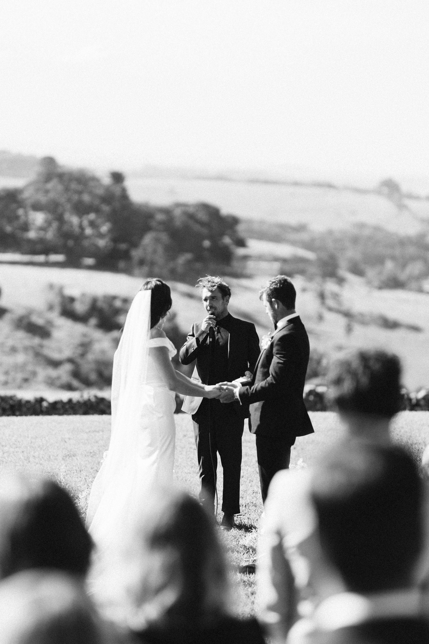 Black and white outdoor wedding ceremony on a hilltop with guests watching.
