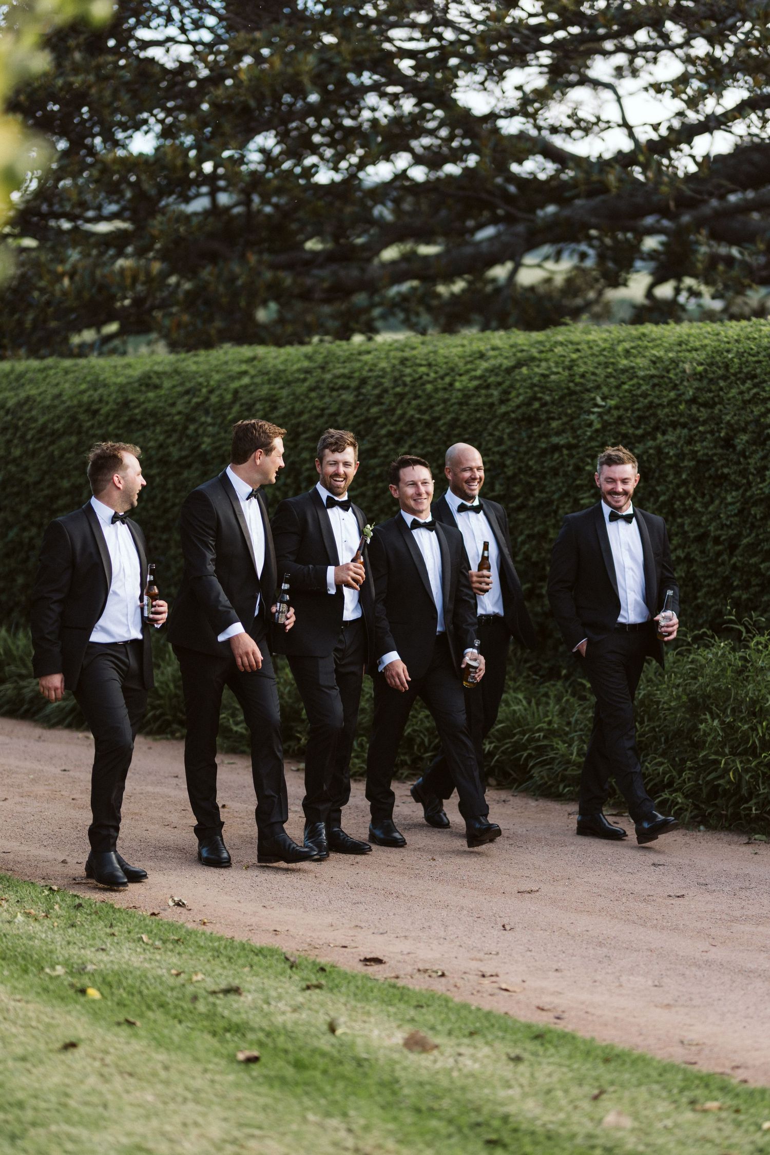 Groomsmen in black tuxedos walking along garden path surrounded by manicured hedges.