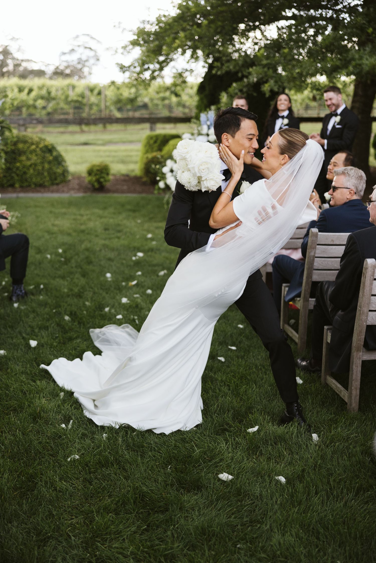 Newlyweds share a romantic dip kiss during outdoor wedding ceremony with flower petals falling around them.