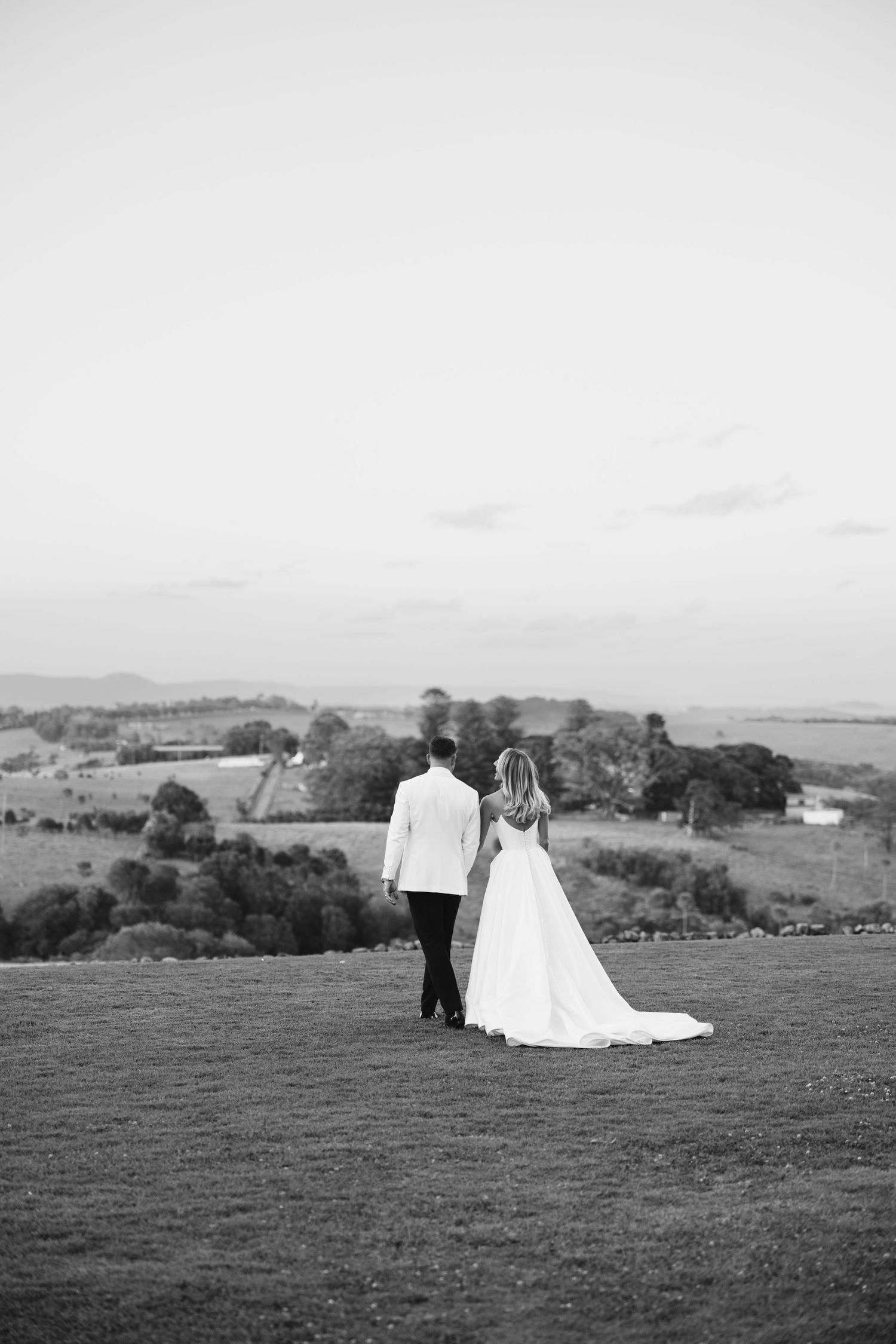 A wedding couple stands on a hilltop overlooking rolling countryside in black and white.