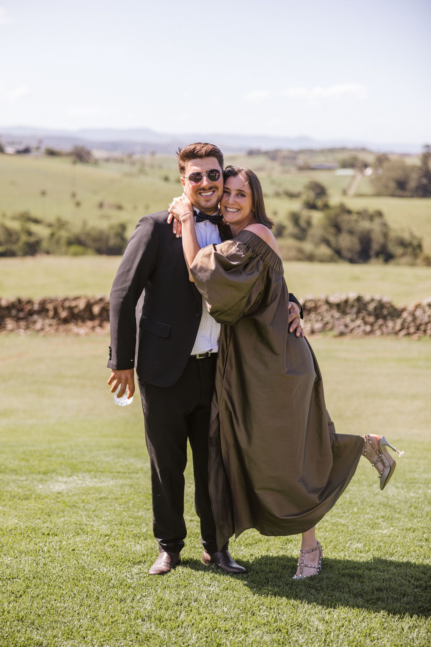 A couple in formal attire poses playfully on a grassy hillside with scenic countryside views.
