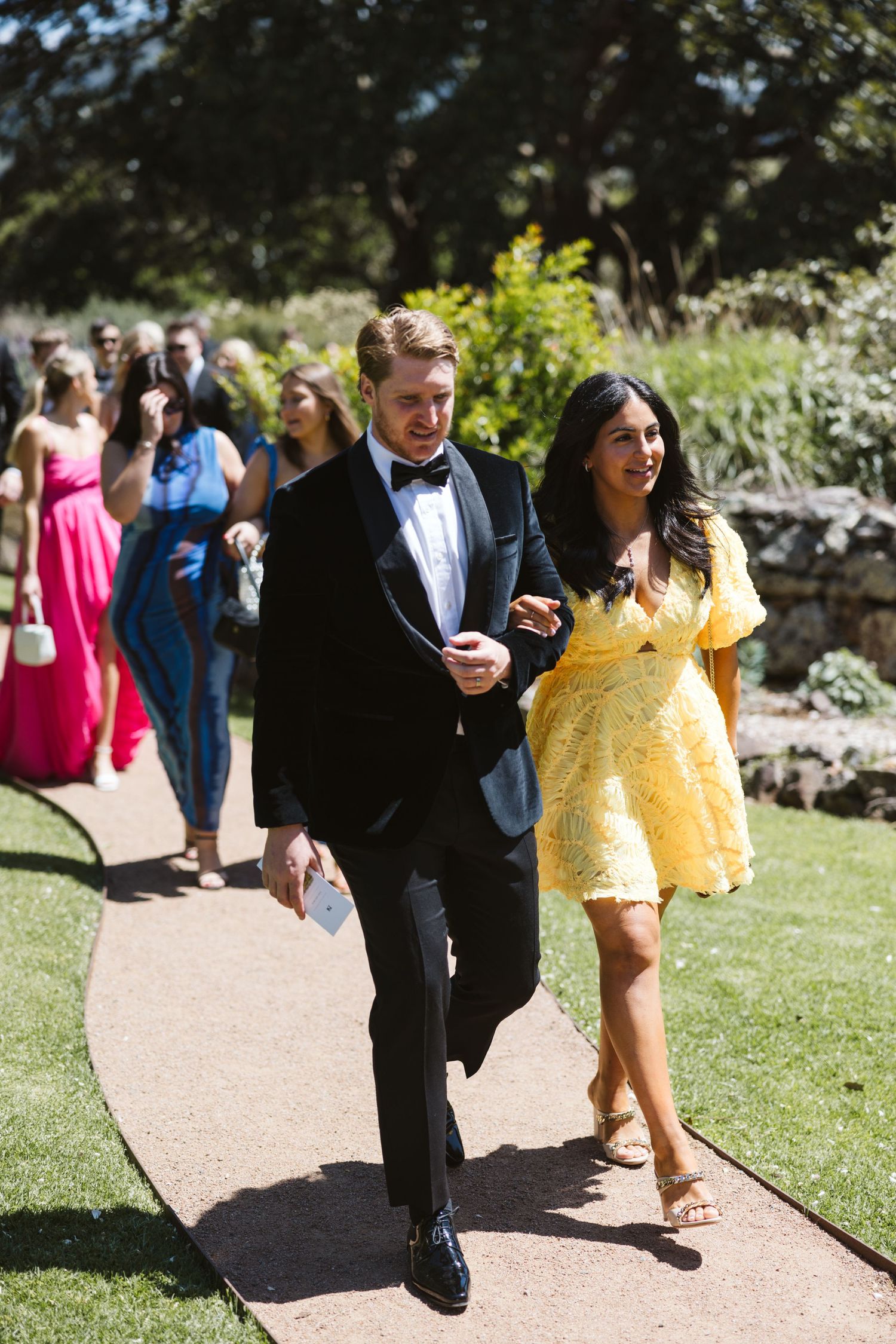 Wedding guests walking along garden path with woman in yellow dress and man in tuxedo.