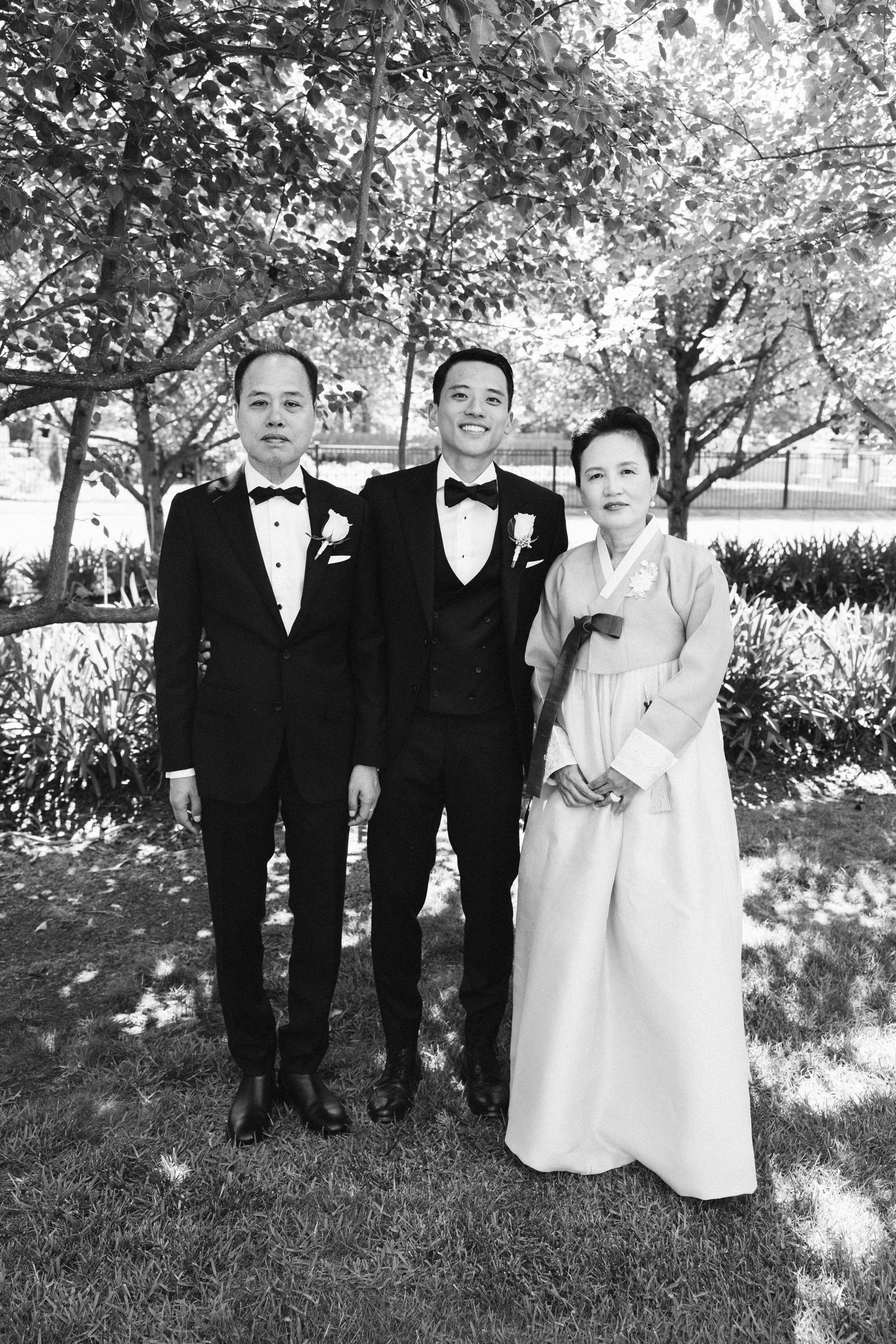 A formal wedding photo outdoors with three guests in traditional black tuxedos and hanbok attire.