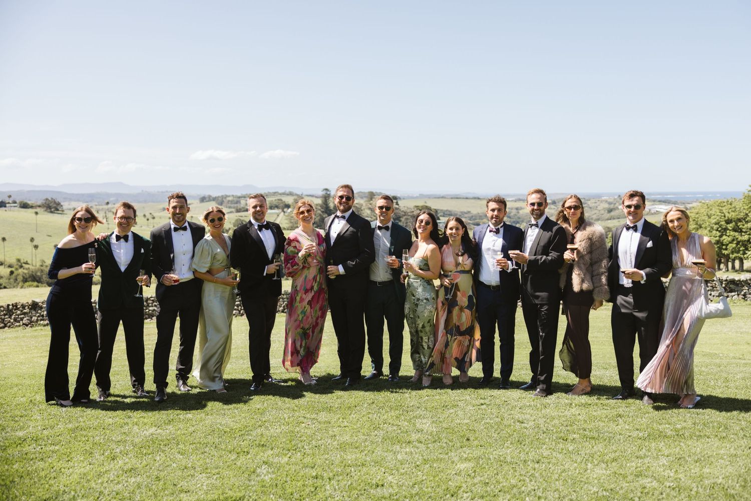 Large wedding party group photo taken on a grassy hillside with scenic views.