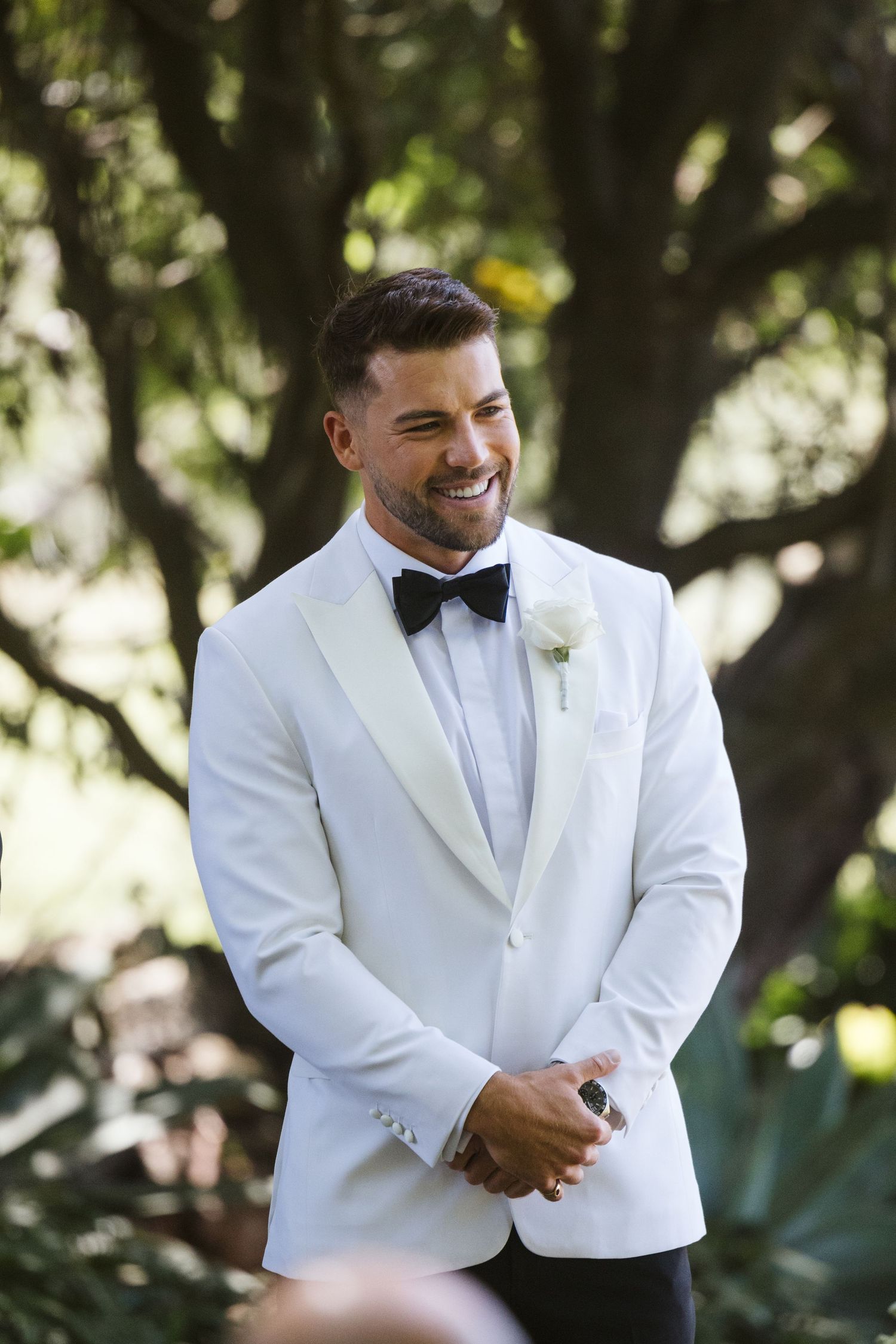 Groom in white dinner jacket smiling during outdoor wedding ceremony.