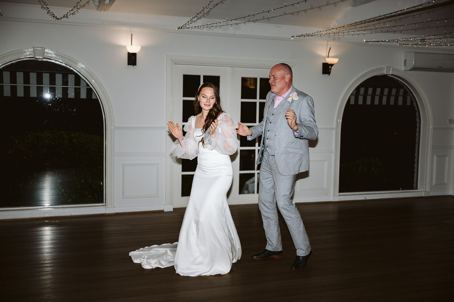 Bride in white gown and groom in gray suit dancing joyfully at elegant wedding reception venue.