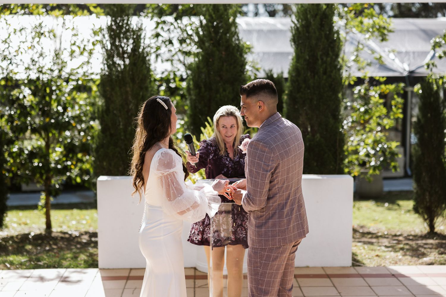 Couple exchanging vows outdoors with officiant at elegant garden wedding ceremony on sunny day.