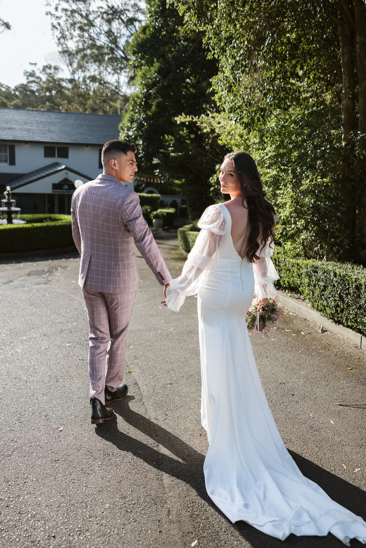 A bride and groom walk hand in hand down a sunlit path surrounded by lush greenery on their wedding day.