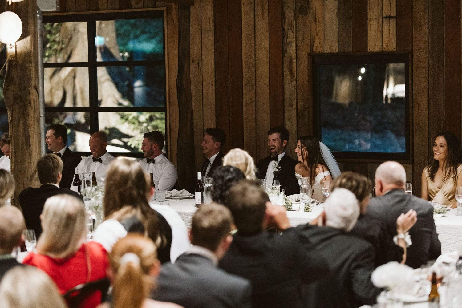 Wedding reception dinner in a rustic wooden venue with guests seated at long tables.