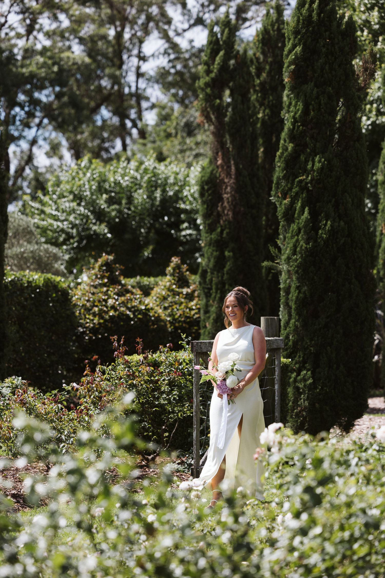 Person in elegant white dress holds bouquet while standing in garden path lined with cypress trees.