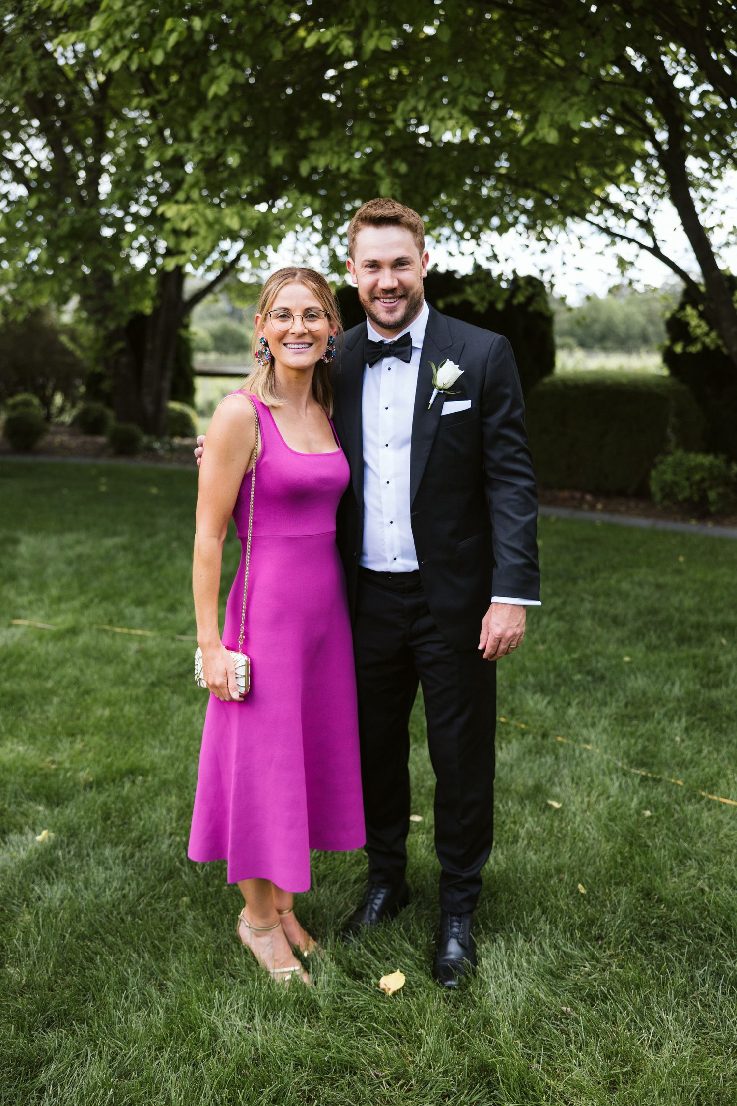 A wedding guest in a magenta dress poses with another guest in formal attire on a lawn.