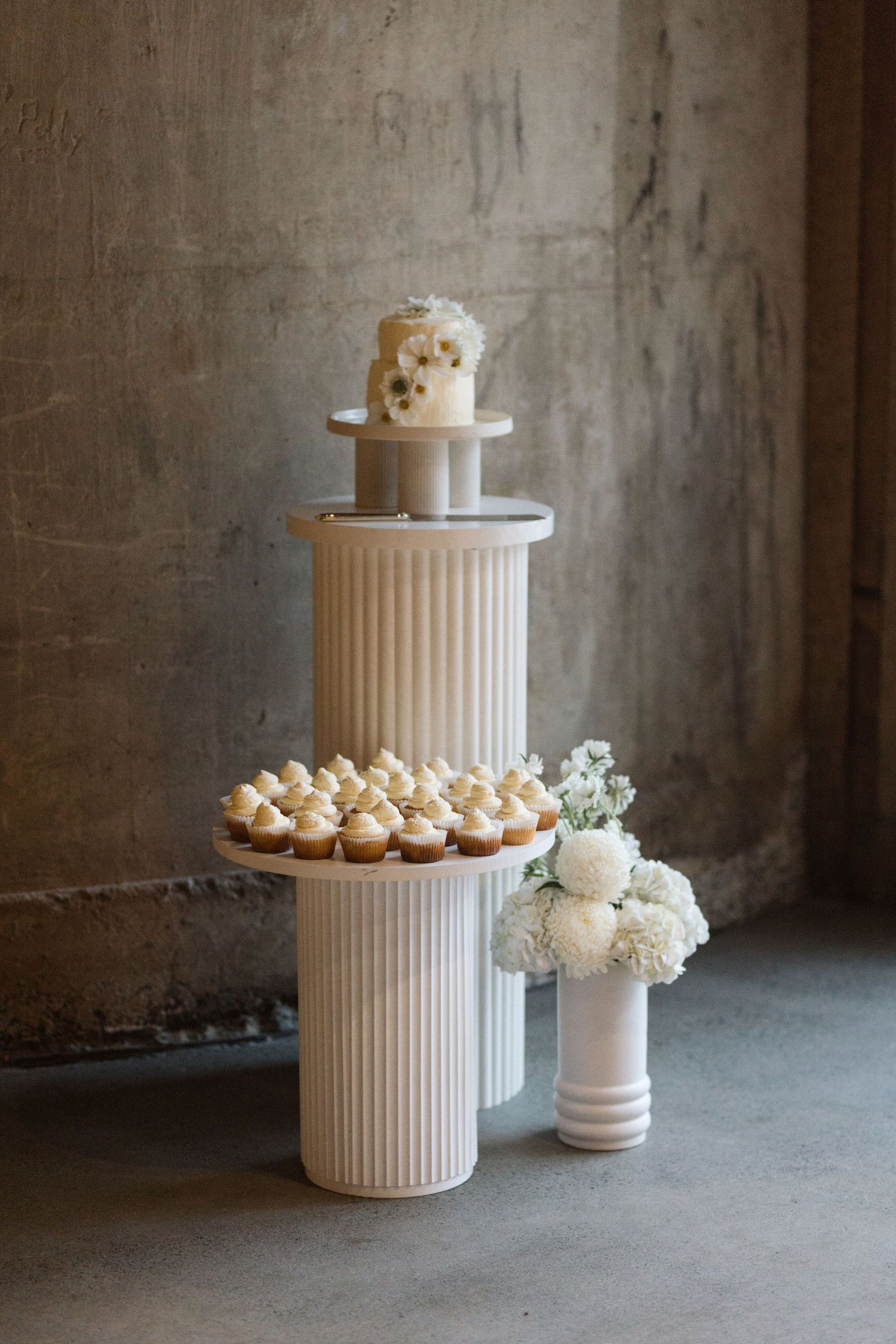 Elegant white wedding cake and cupcakes displayed on modern fluted pedestals with fresh flowers.