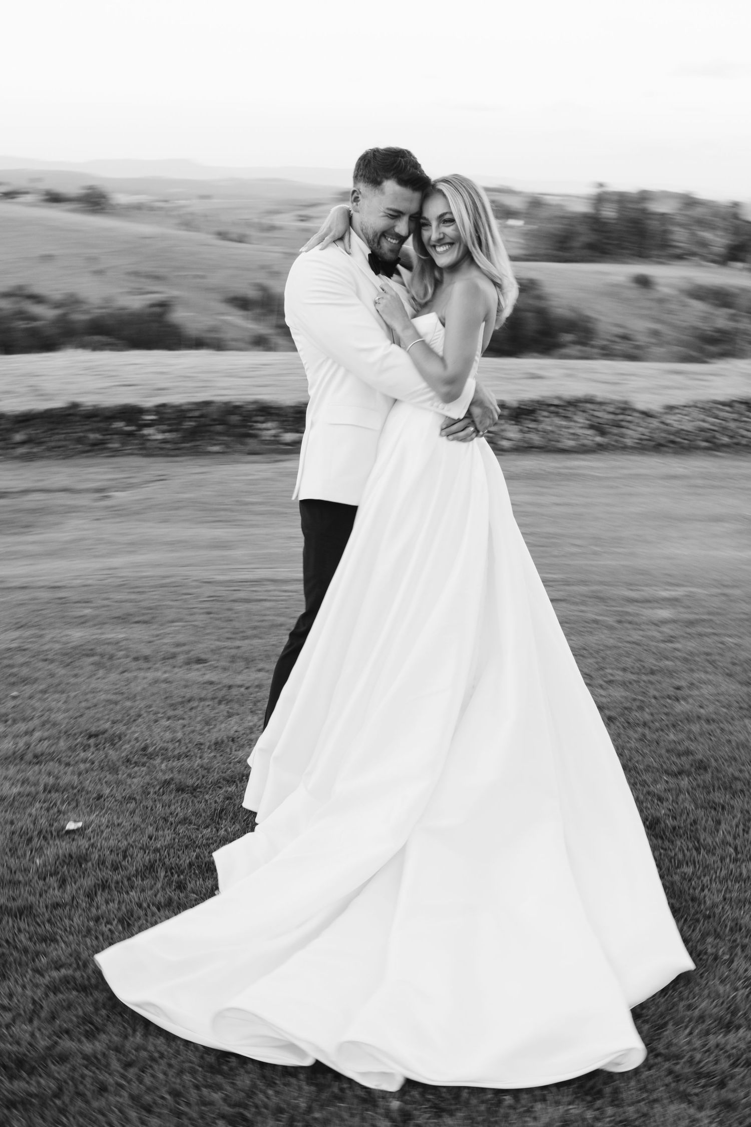 A black and white photo of newlyweds embracing on a grassy hillside.