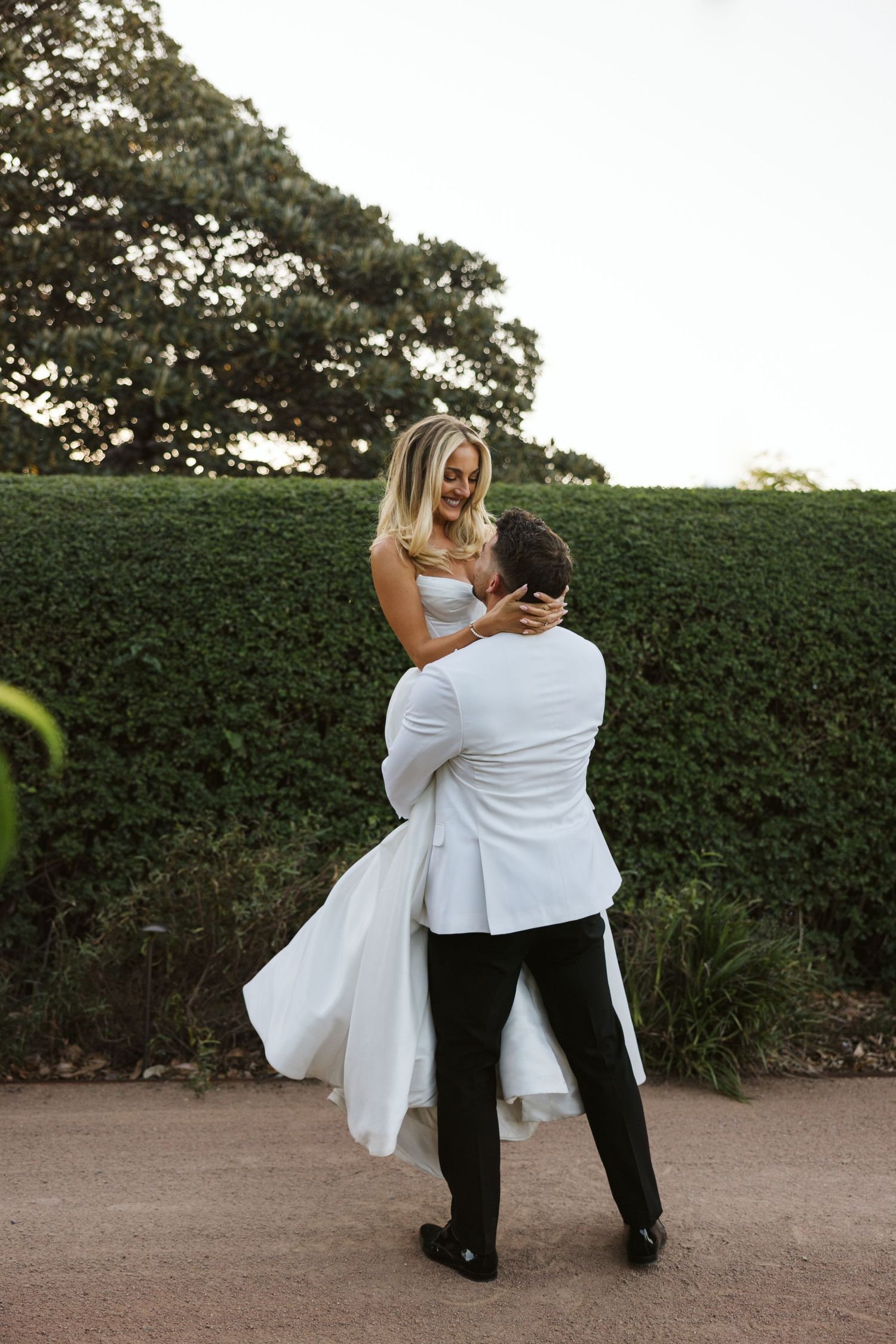 Groom lifts bride in elegant white dress against garden hedge backdrop.