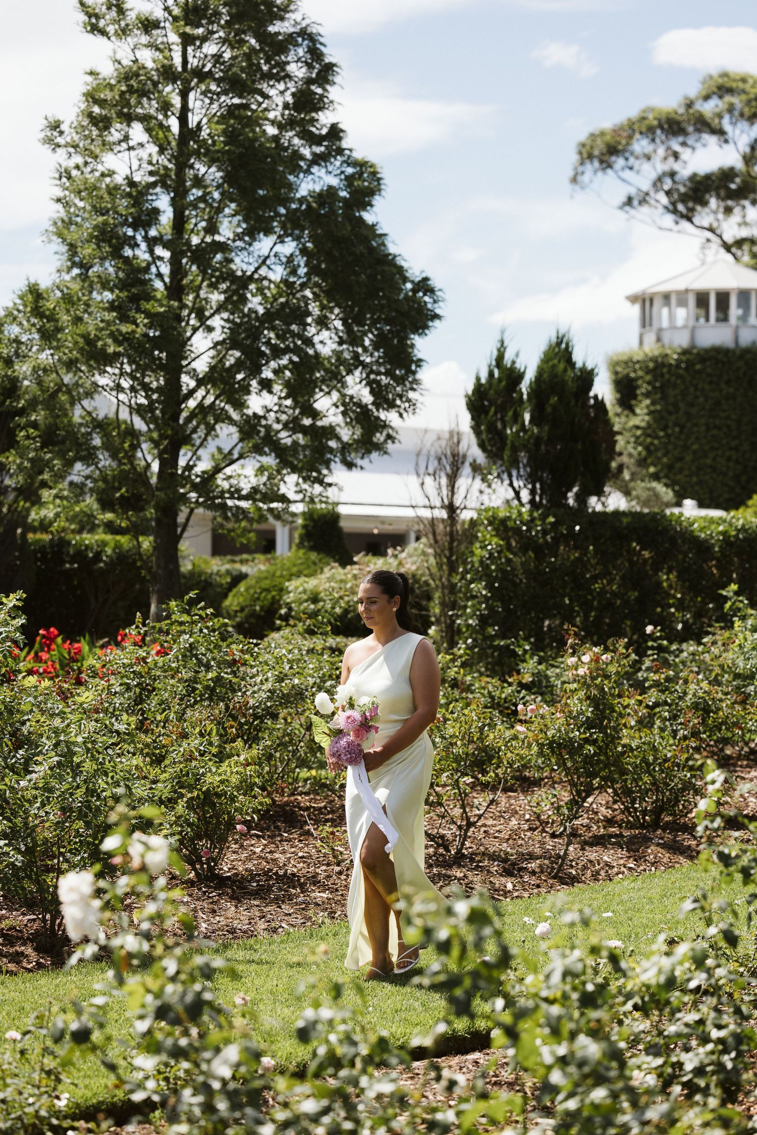 Person in sleek white dress holds bouquet while standing in blooming garden with white building in background.