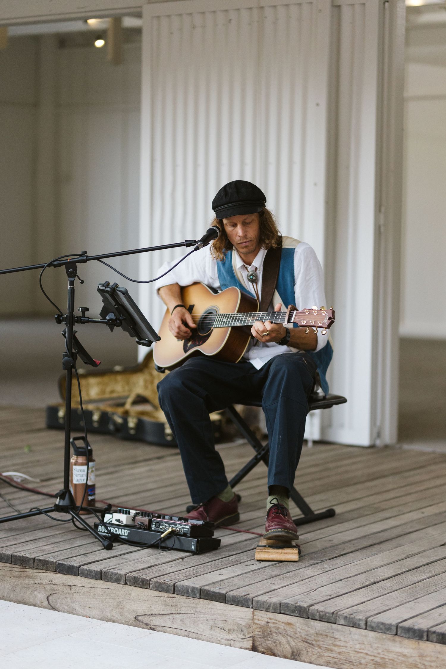 A solo musician plays acoustic guitar while seated on stage with a music stand and effects pedals.