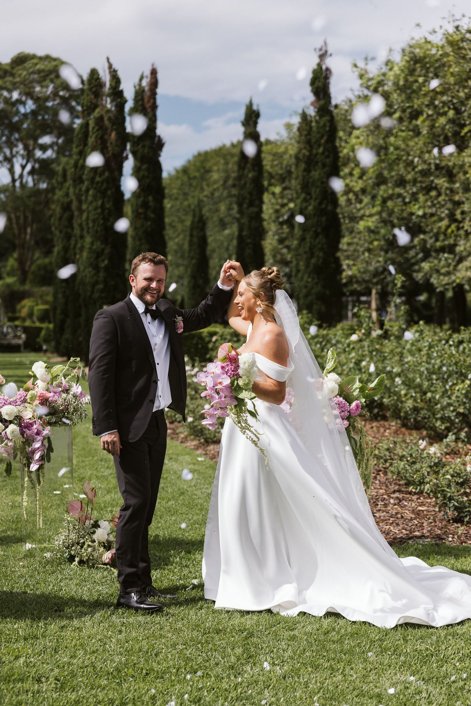 Elegant garden wedding scene with tall cypress trees and pink roses as couple poses on manicured lawn.