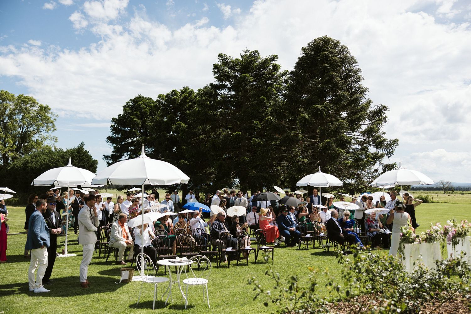 Wedding guests mingling under white umbrellas at outdoor garden reception.