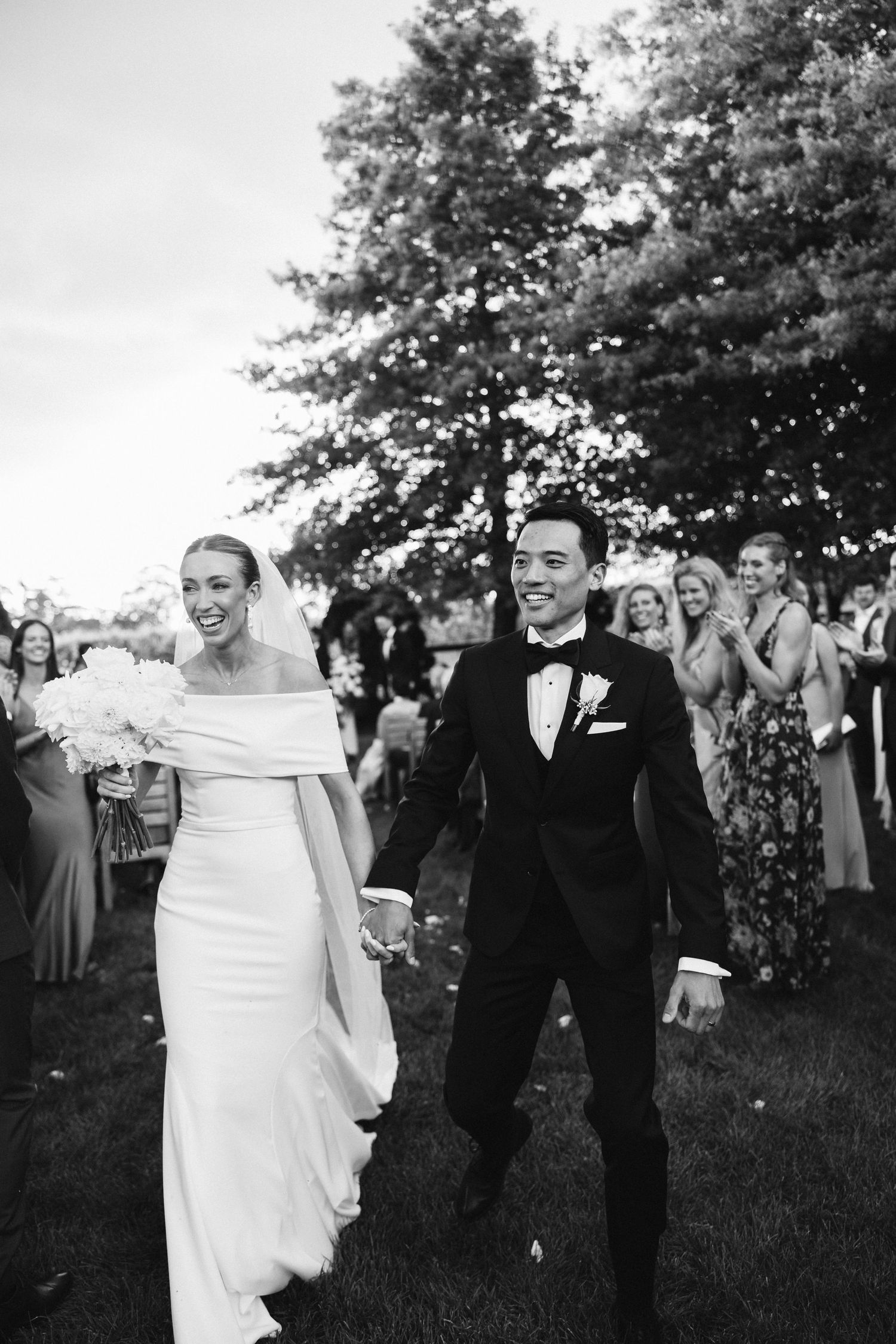 Black and white photo of joyful newlyweds walking down aisle at outdoor garden ceremony.