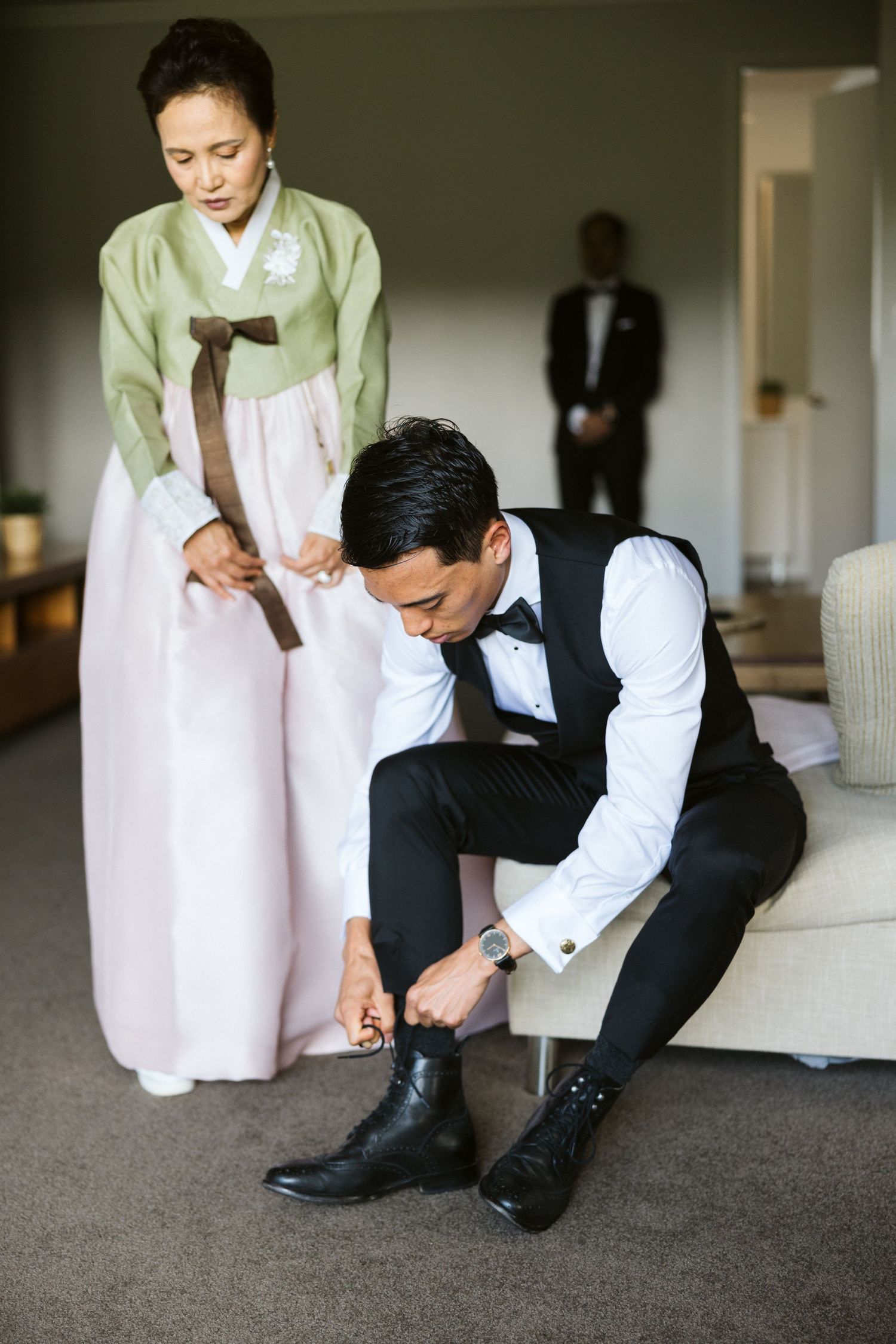 A groom in formal attire ties his shoes while someone in traditional Korean hanbok stands nearby.