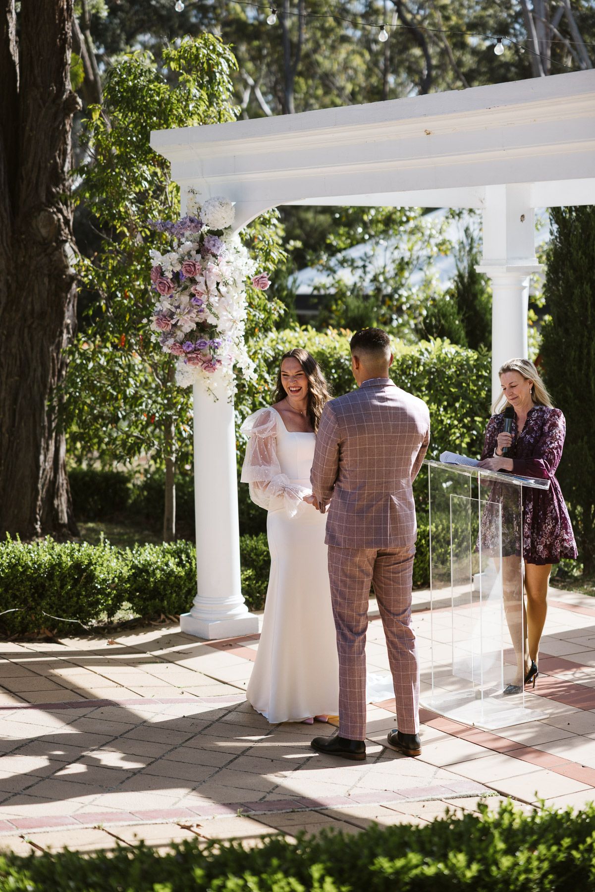 Couple exchanging vows at an outdoor wedding ceremony under a floral pergola, officiated by a smiling woman at a podium.