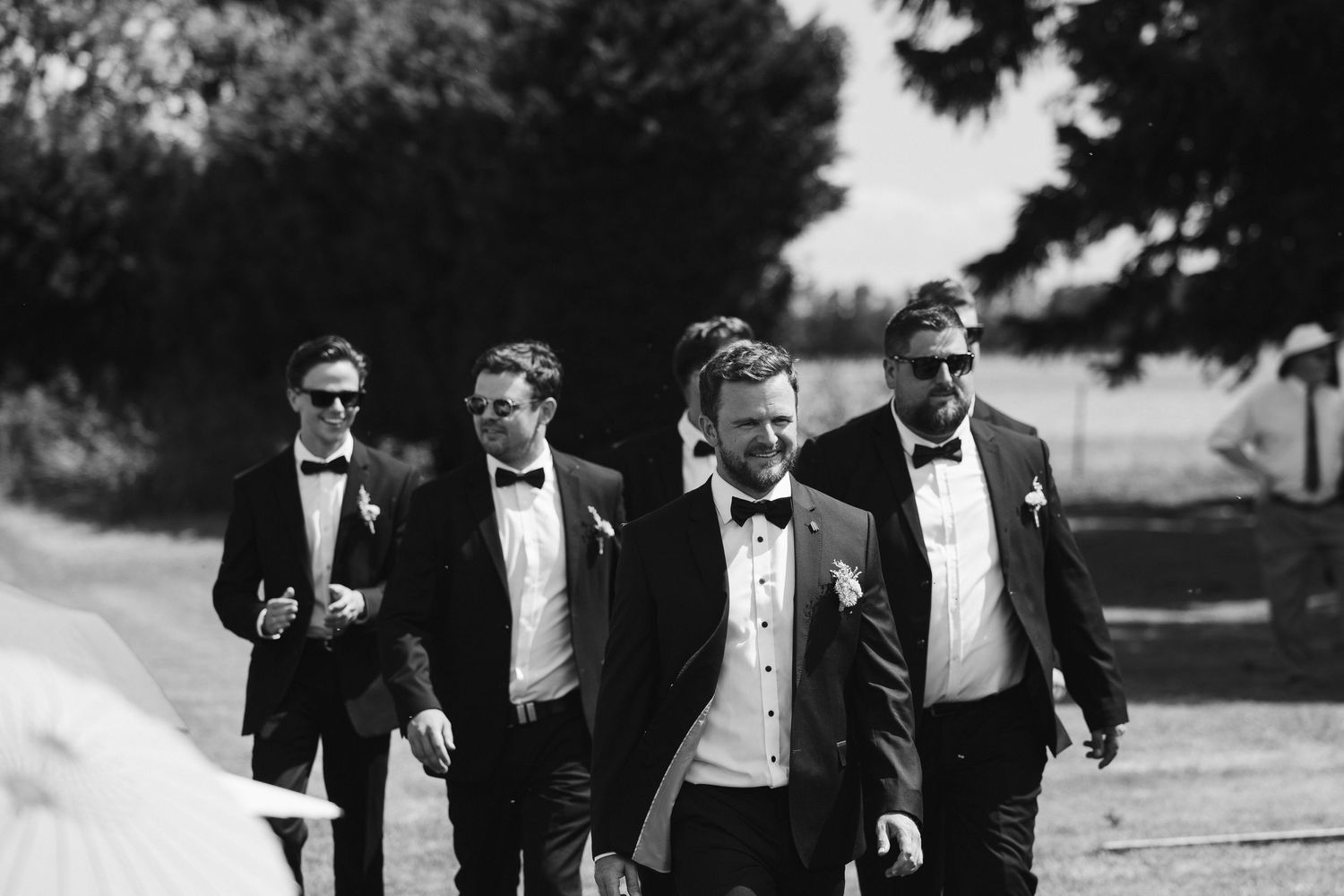 Four groomsmen in black tuxedos and bowties walk together on a path in this black and white wedding photo.