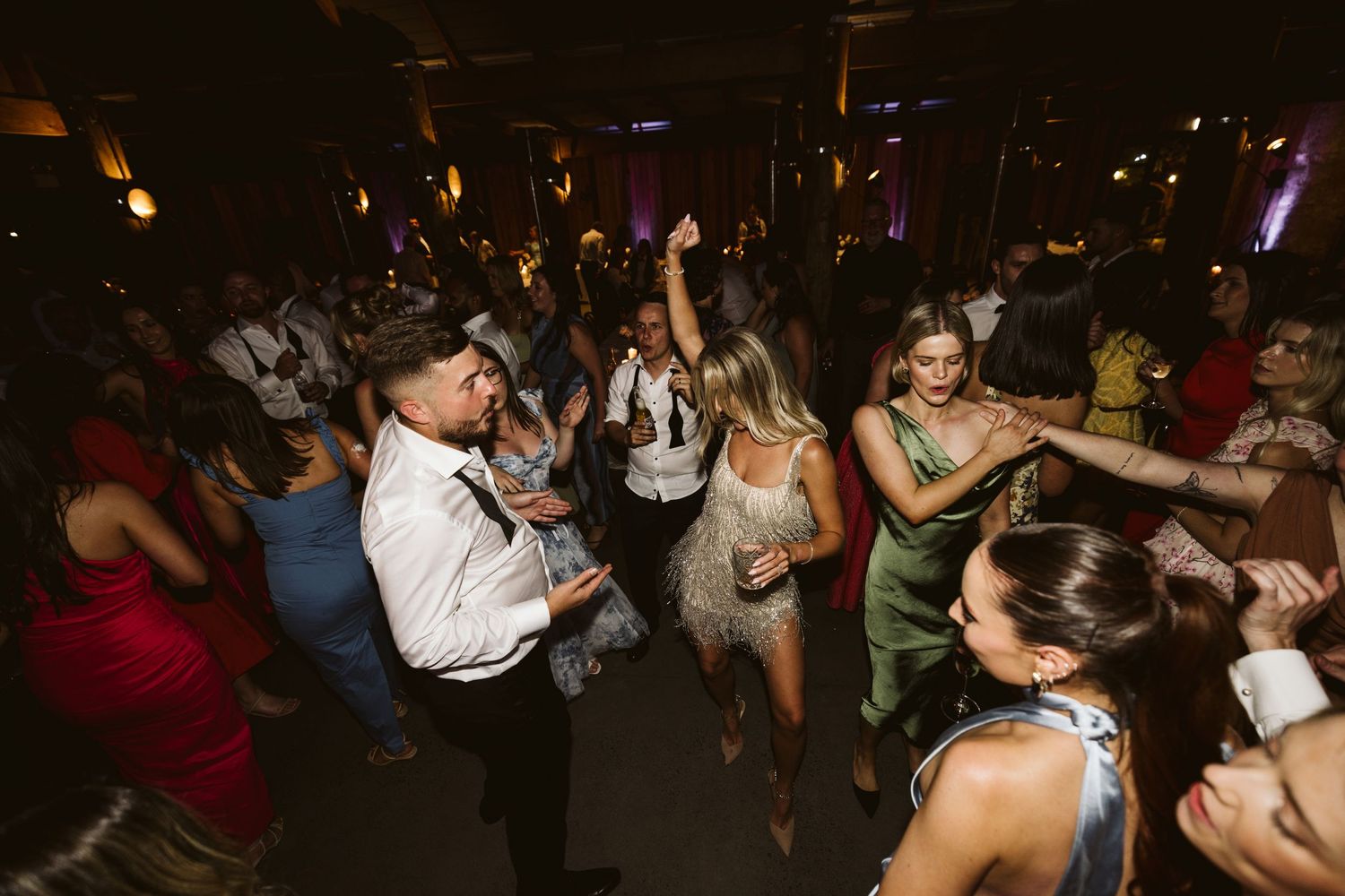 Wedding guests dancing enthusiastically together on a dark dance floor during a reception celebration.