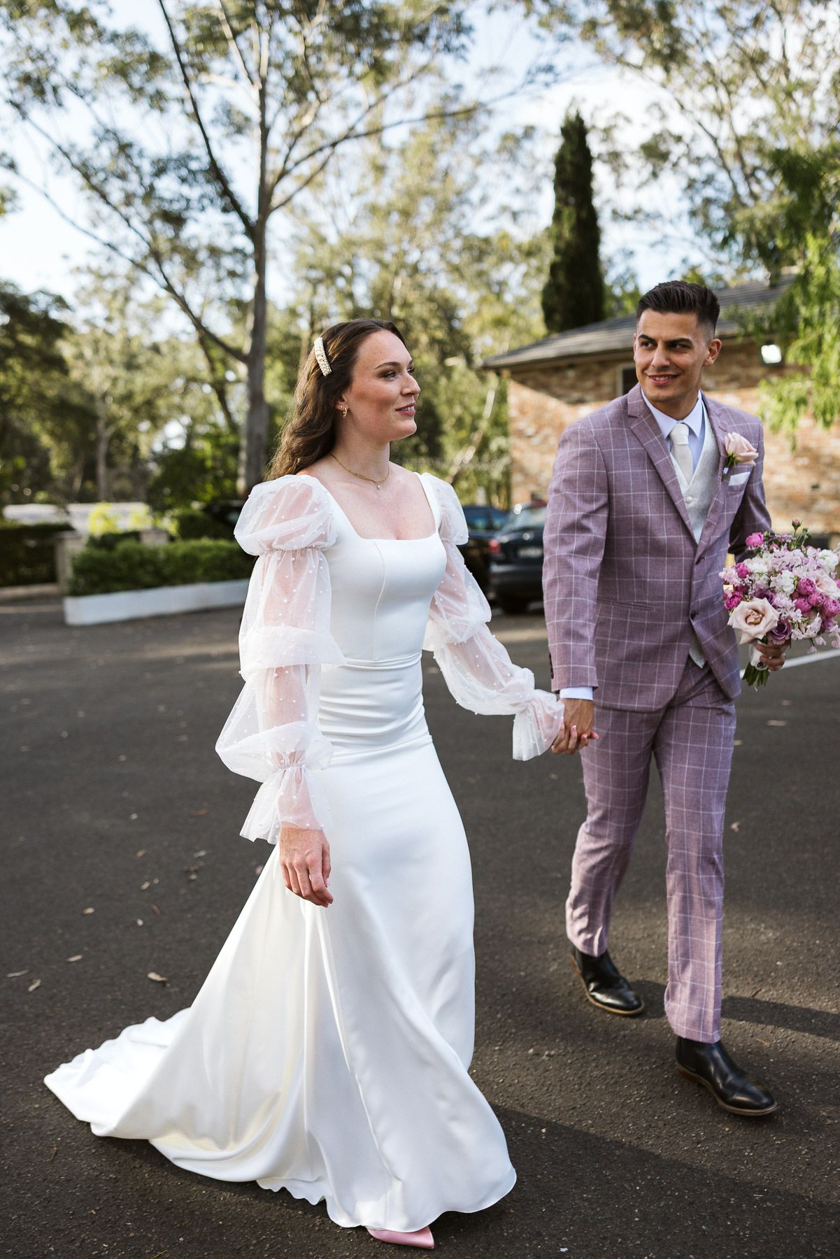 Bride in white gown with sheer sleeves walks hand-in-hand with groom in pink plaid suit holding bouquet outdoors.