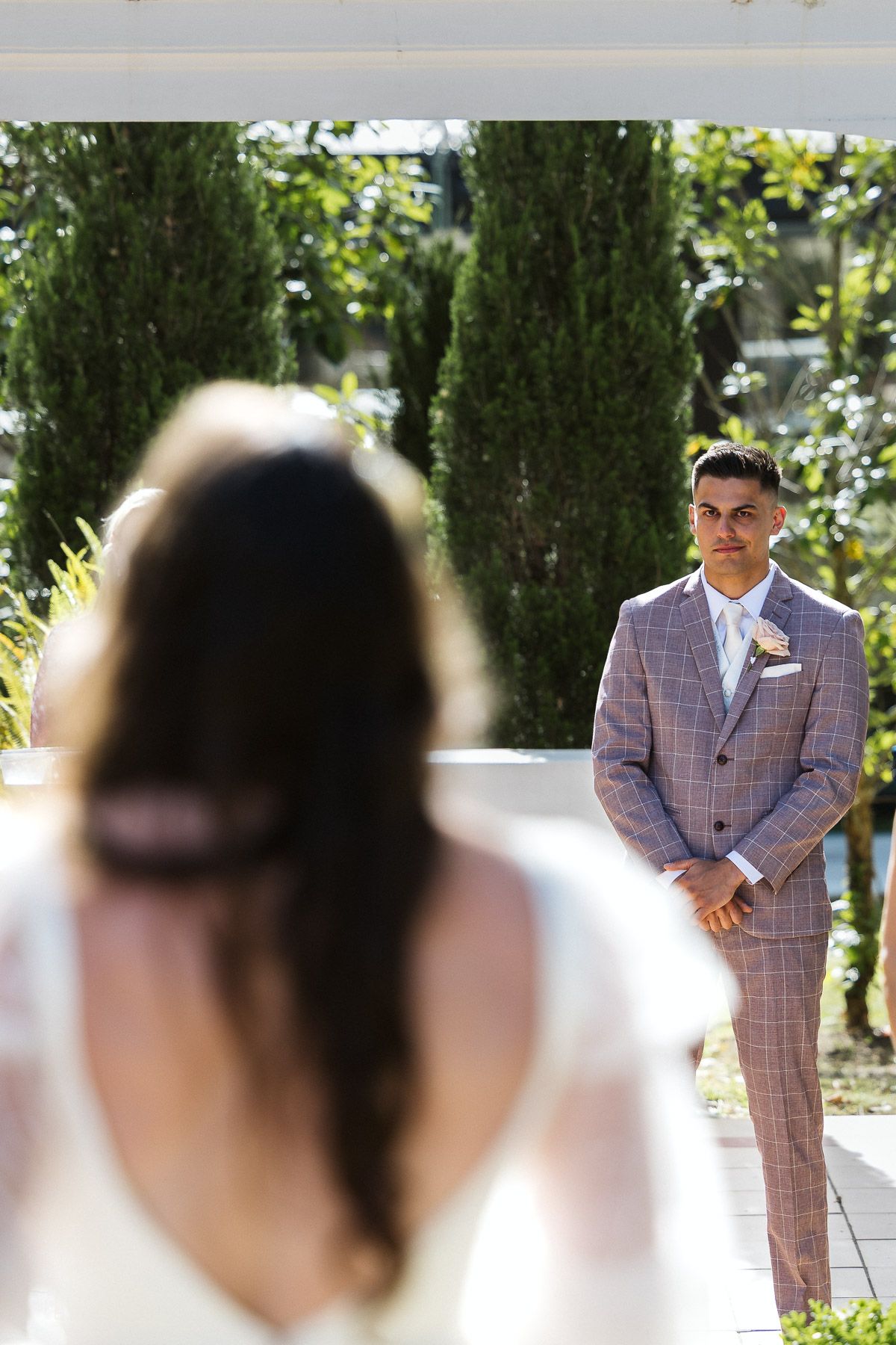 Groom in plaid suit watches bride walk down aisle at outdoor wedding ceremony surrounded by cypress trees.