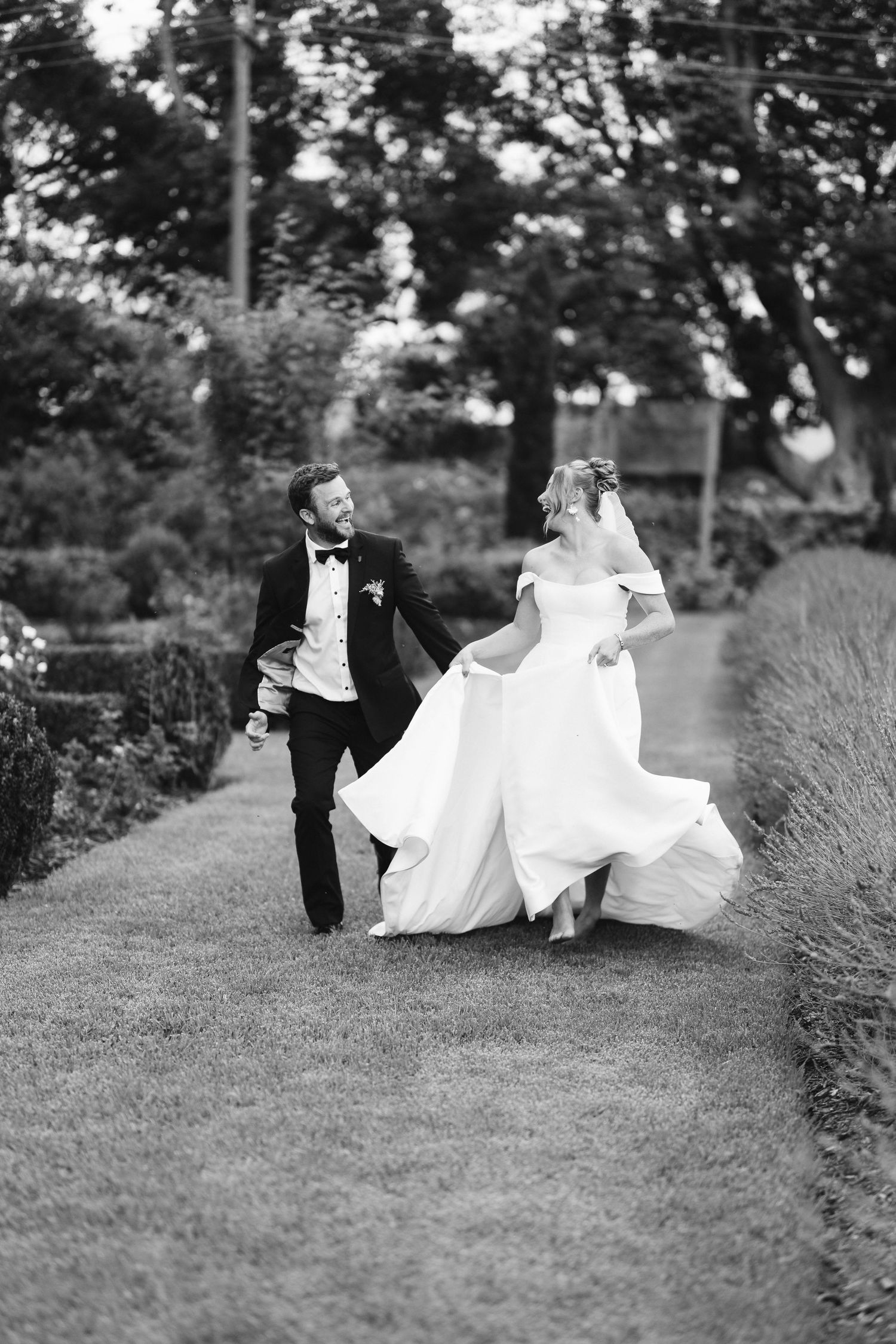 Black and white photo of newlyweds running playfully along garden path with bride's dress flowing.