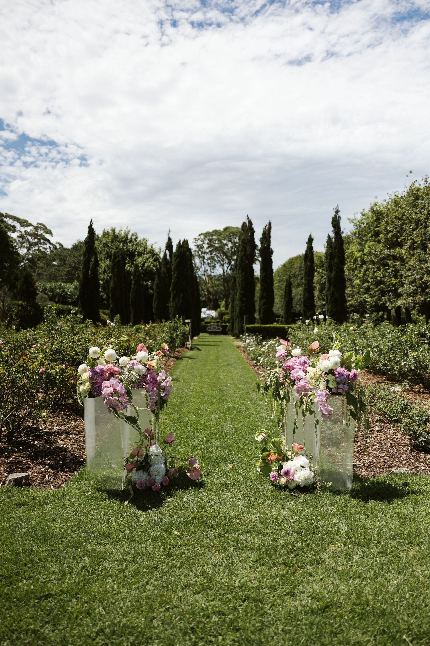 Outdoor wedding aisle decorated with tall glass vases of pink flowers between manicured gardens and cypress trees.