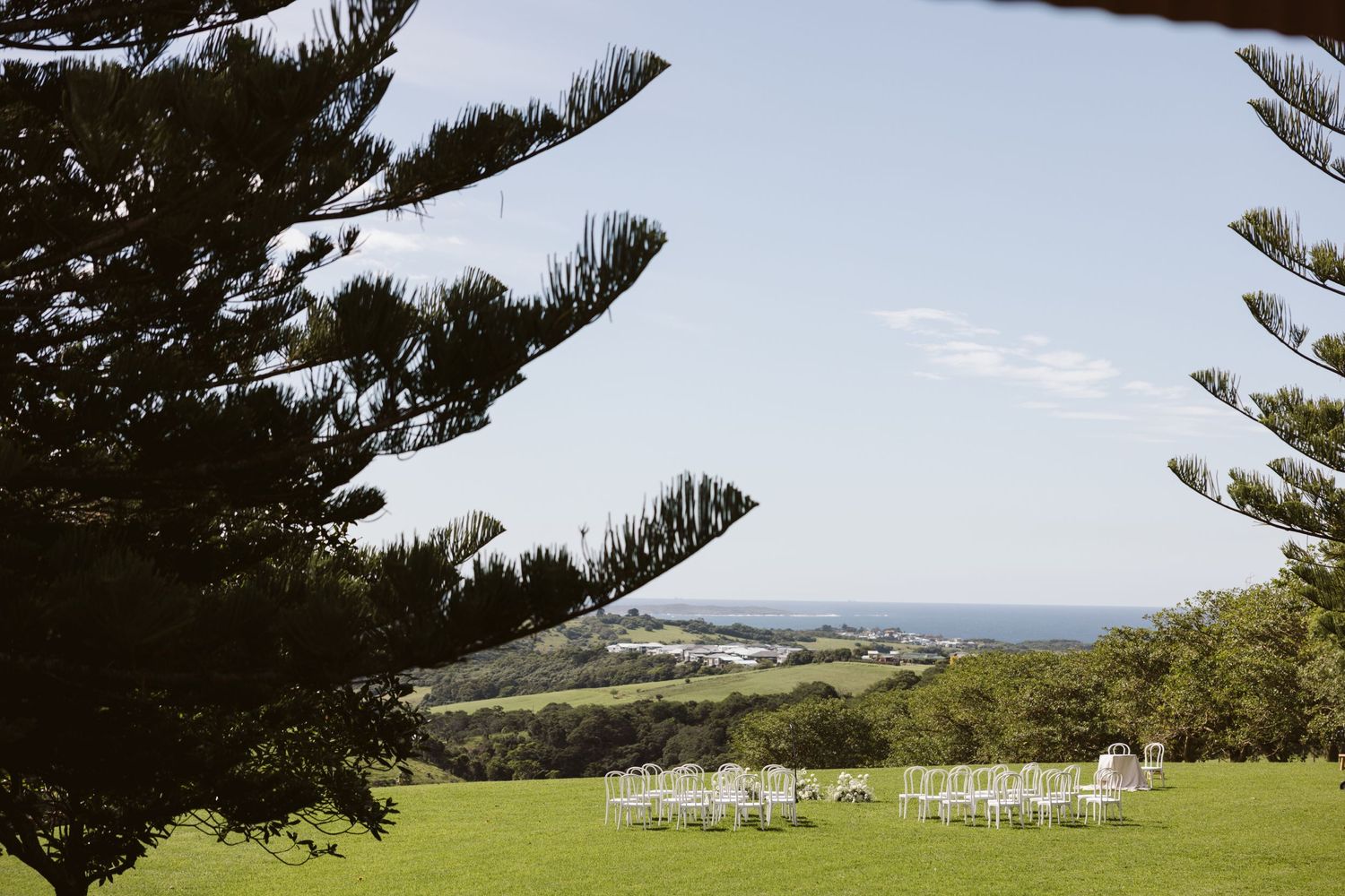 White ceremony chairs set up on hilltop lawn framed by towering pine trees with ocean view.