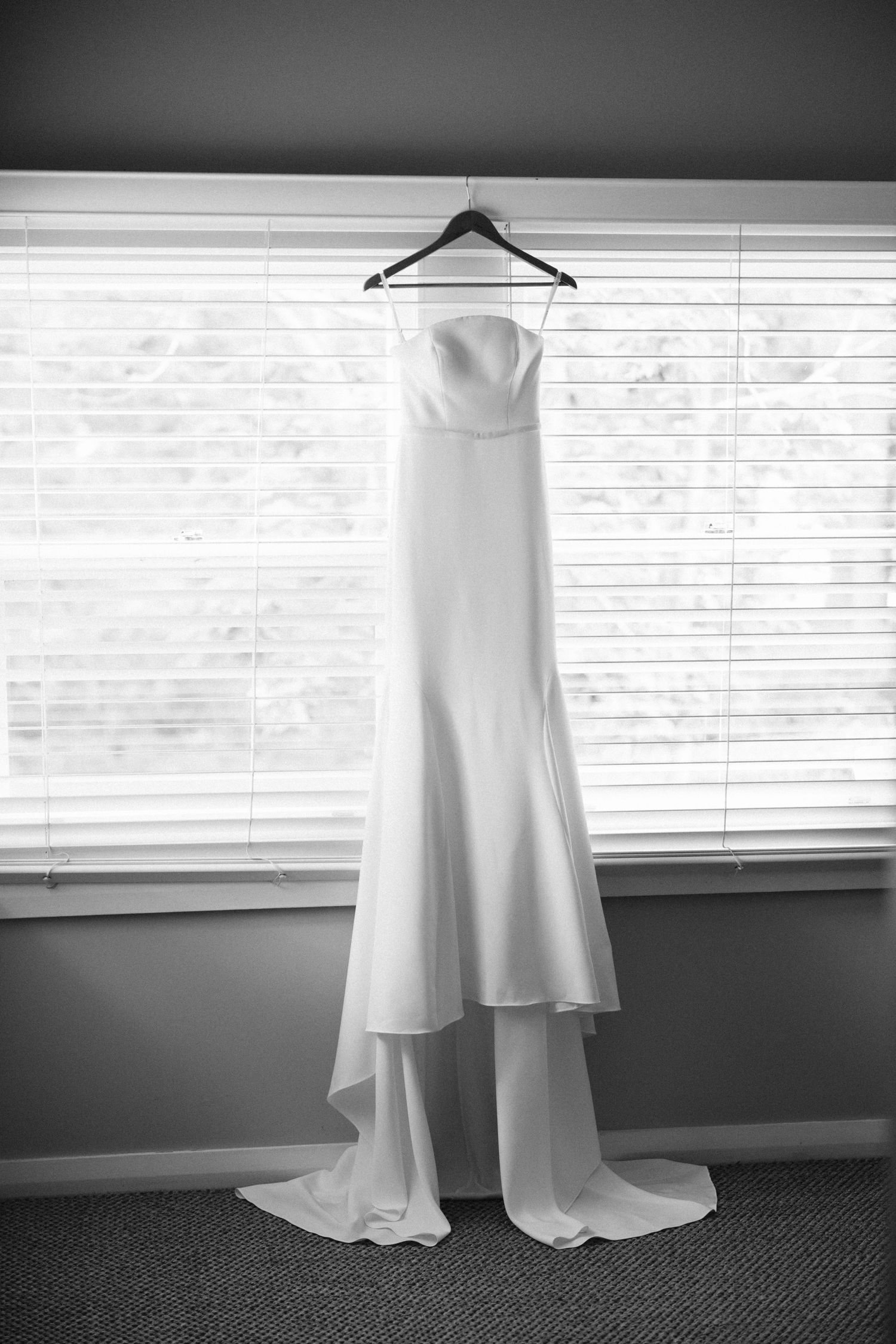 A white wedding dress hanging on display against venetian blinds in black and white.
