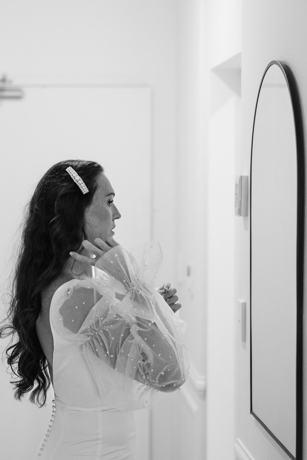 Black and white photo of a bride in a white robe adjusting her earring while looking in a mirror.