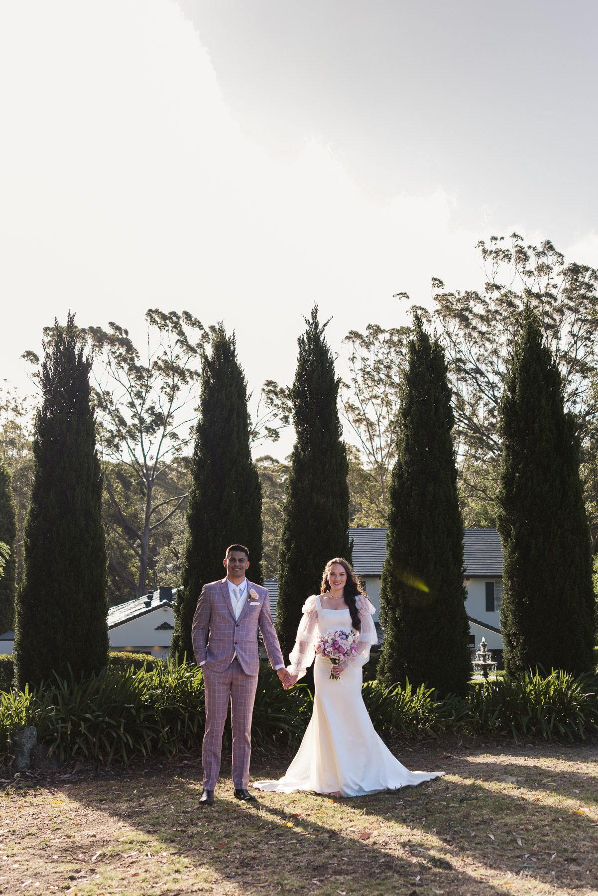 Bride and groom holding hands in garden with tall cypress trees, bride in white gown holding purple bouquet, groom in pink suit.
