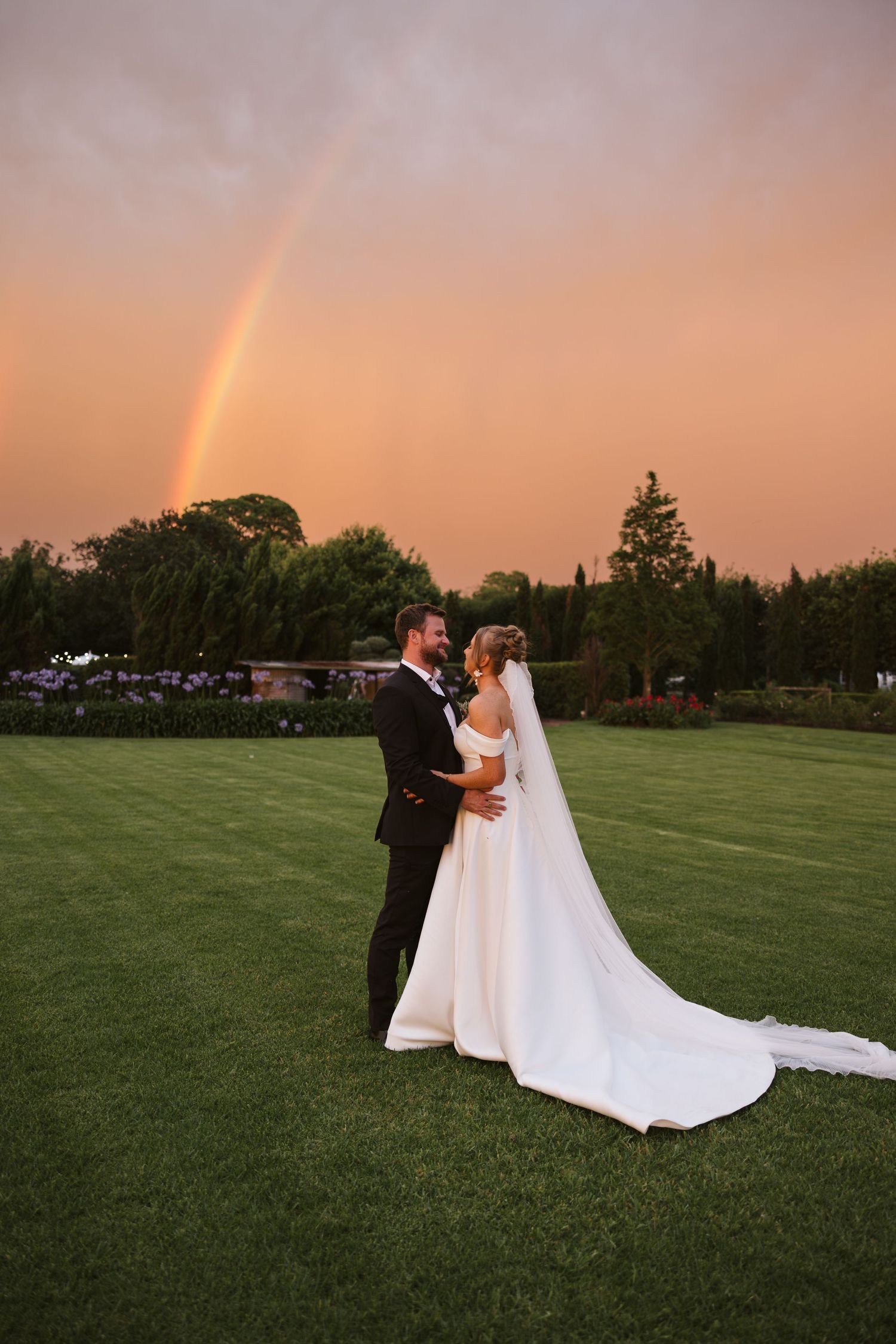 Newlyweds pose together on grass field with stunning rainbow and sunset behind them.